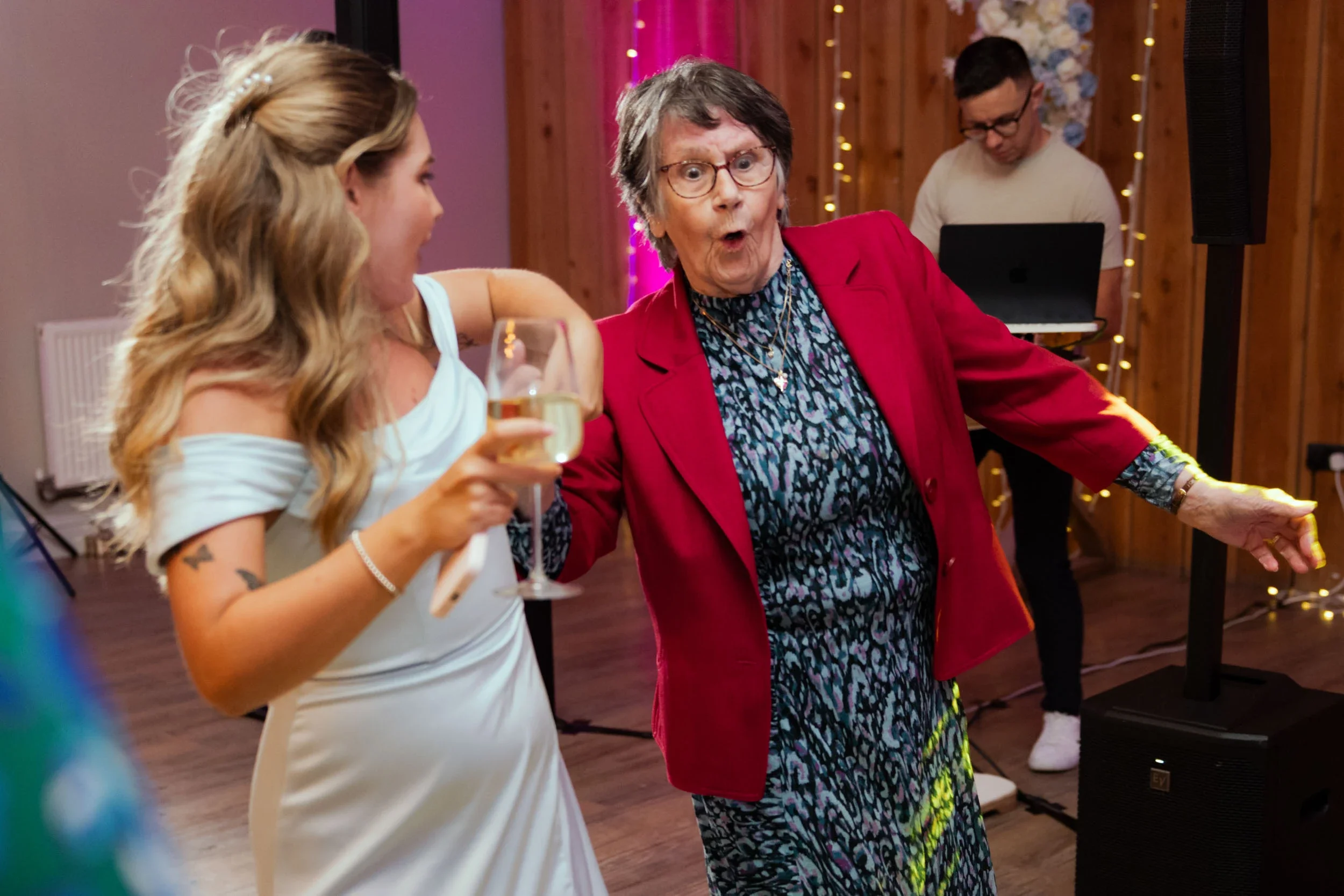 An elderly woman and a young woman dancing at a celebration; the elderly woman wearing a red blazer and patterned dress, showing an excited expression, while the young woman in a white dress holds a glass of champagne and smiles. A man with glasses a