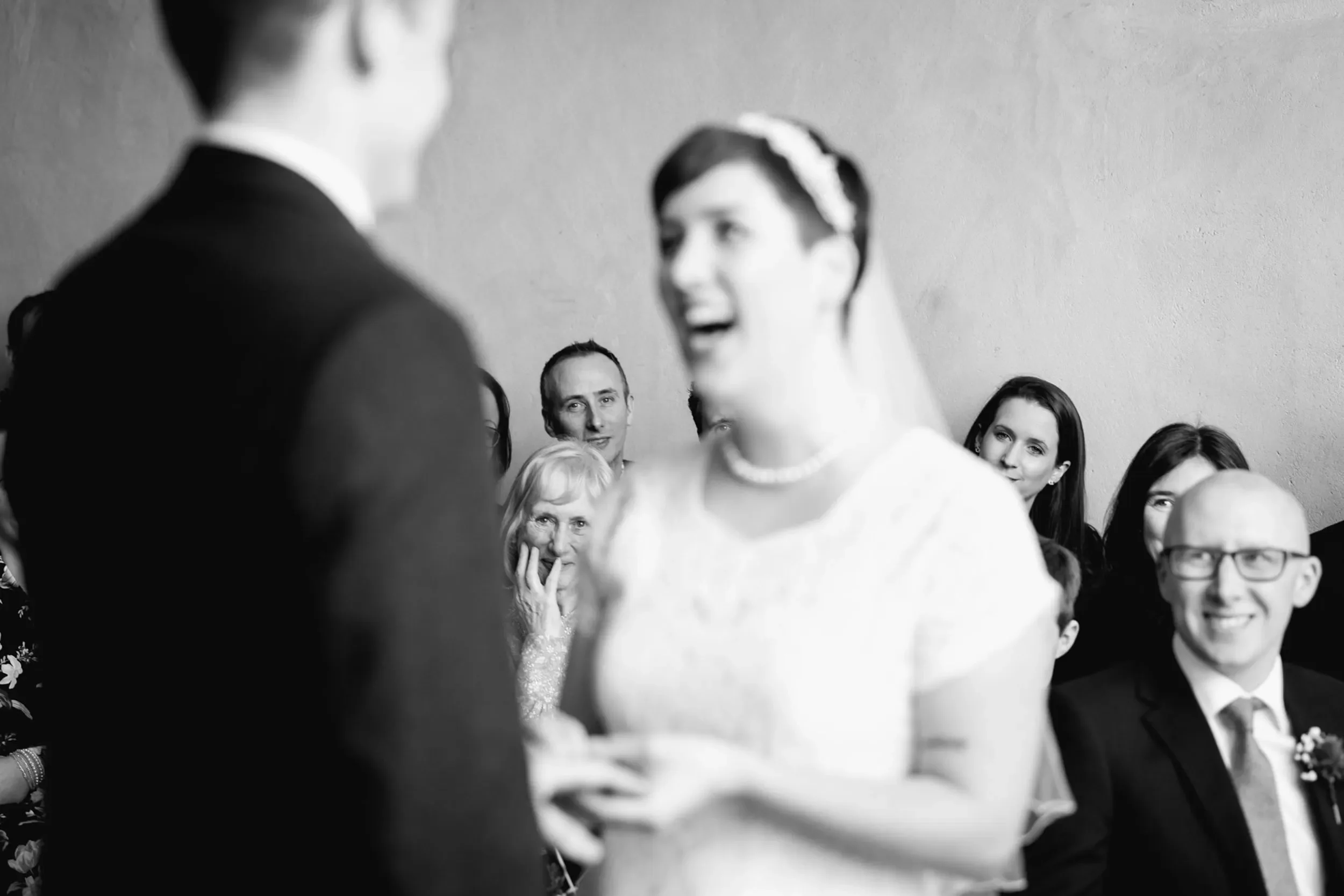 Black and white photo of a wedding ceremony with the bride and groom exchanging vows, surrounded by guests watching with smiles and expressions of joy.