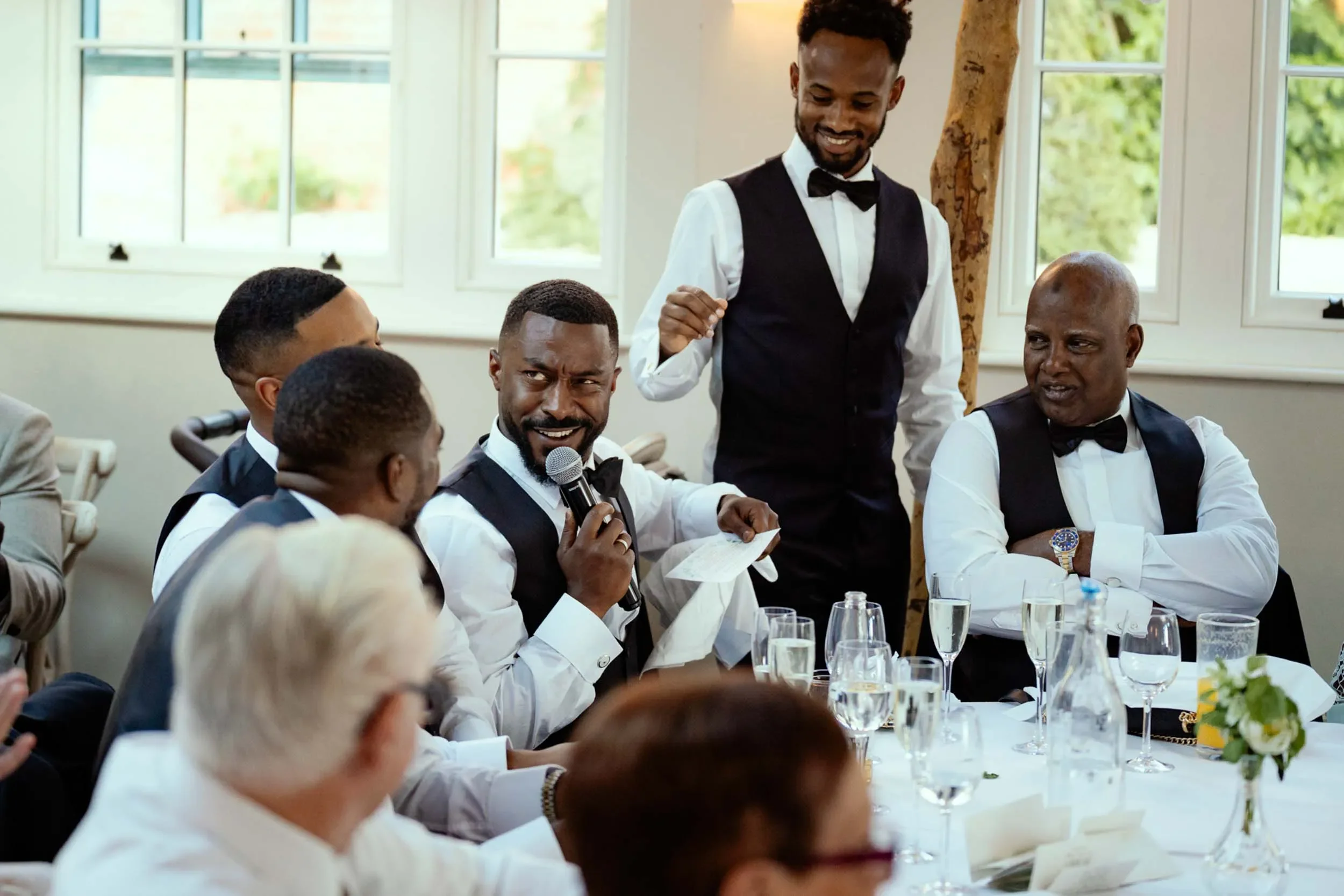 A group of men in formal attire, some with glasses and bow ties, seated around a dining table during a celebration or event, with one man holding a microphone and speaking.