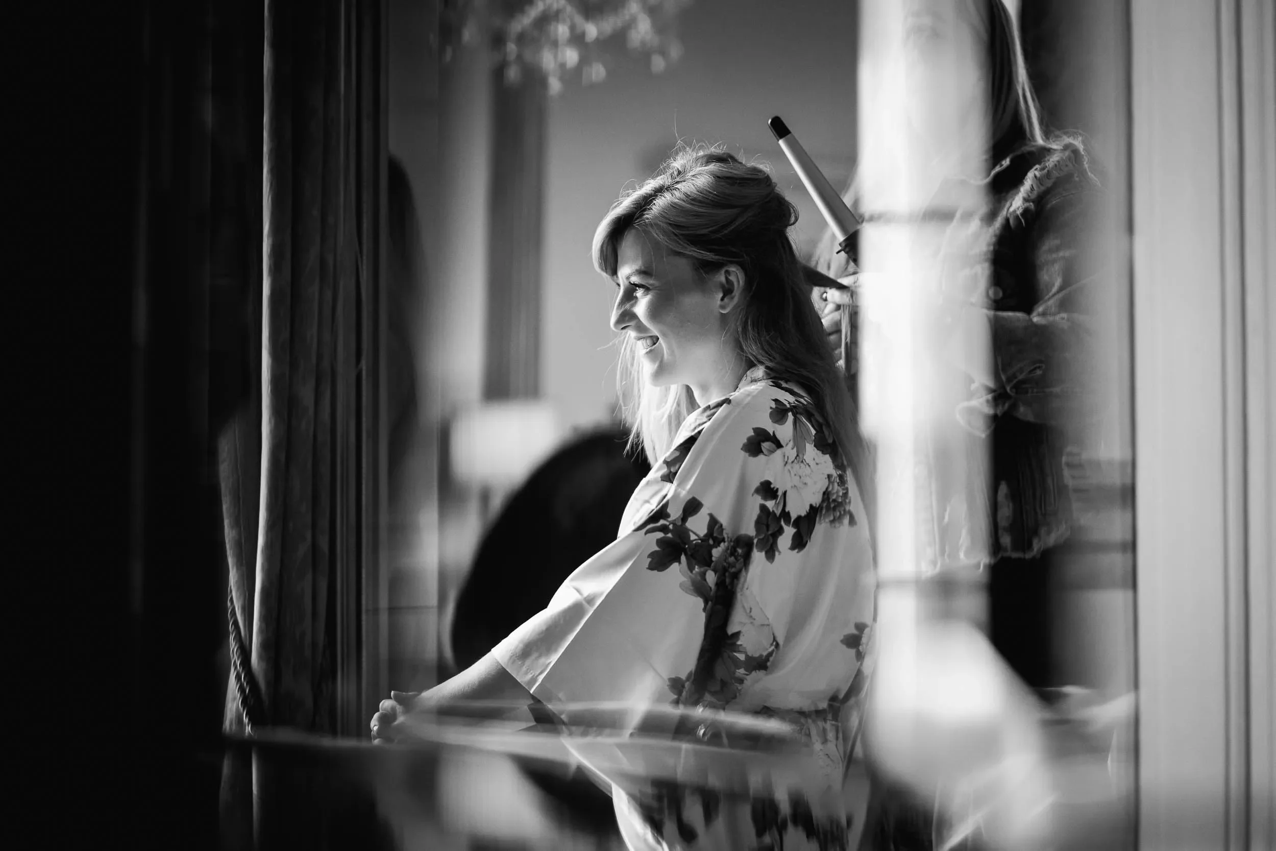 A woman sitting in front of a mirror during a hairstyling session, smiling as a hairstylist works on her hair.