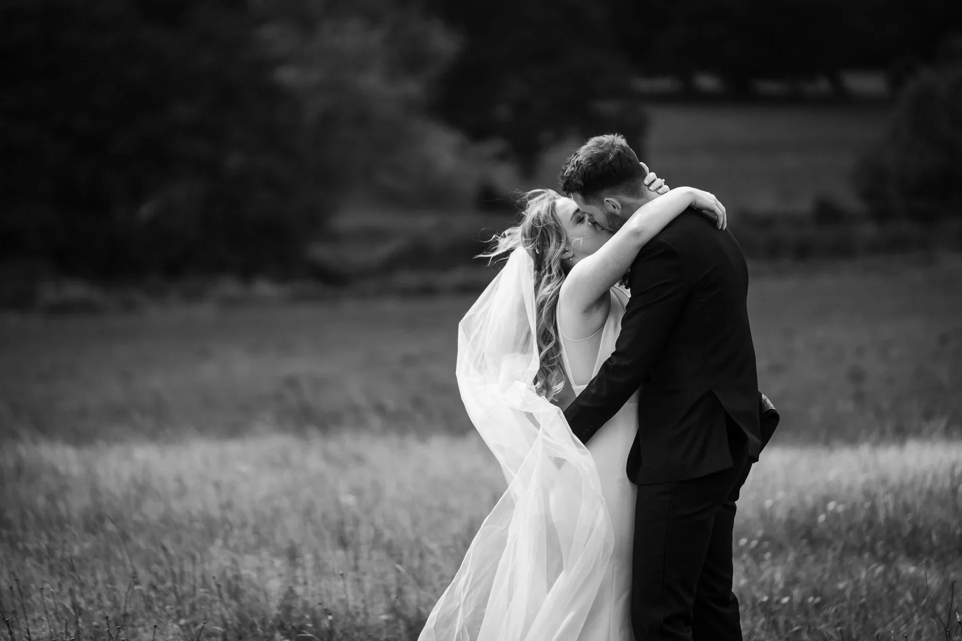 A bride and groom embrace and kiss in an outdoor field, with the bride wearing a wedding dress and veil, and the groom in a dark suit, in black and white photography.