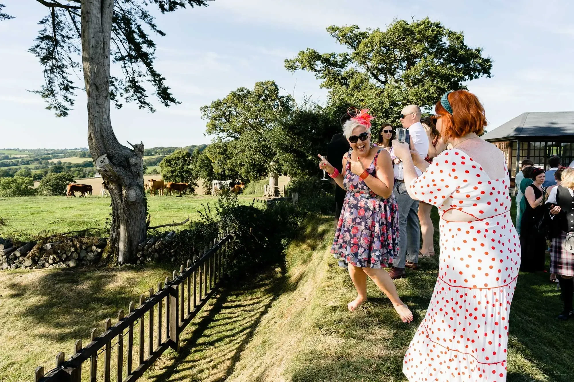 A group of people, mostly women, dressed in vintage and polka dot clothing, stand outside near a fence on a sunny day. They are taking pictures of cows grazing in a field and seem to be enjoying themselves, smiling and posing.