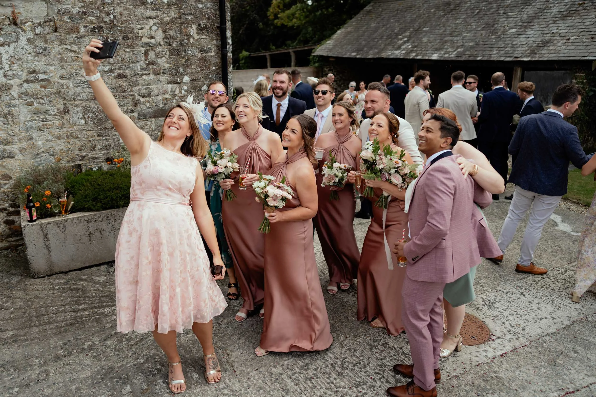 A group of people dressed in formal attire taking a selfie at an outdoor wedding celebration.