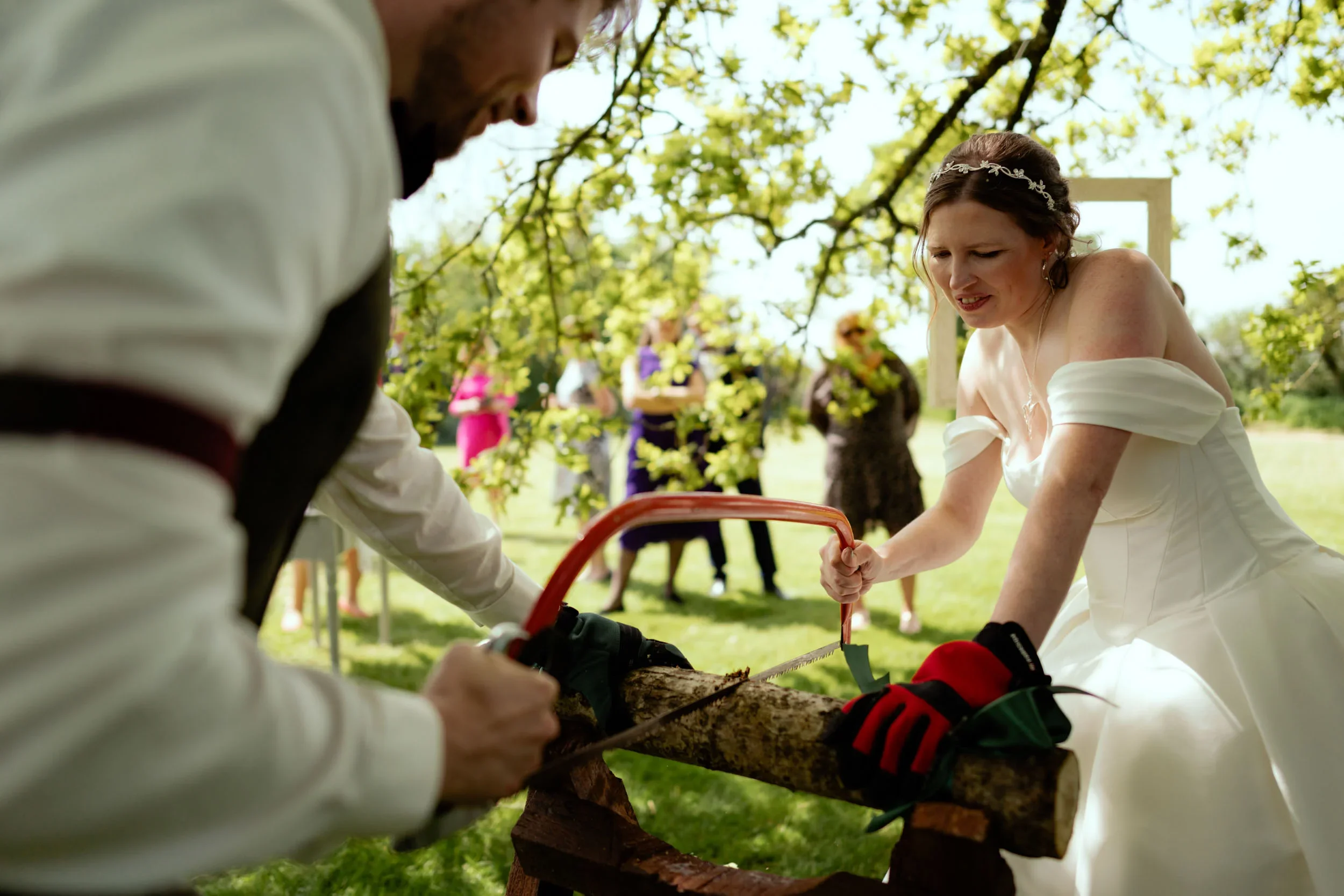 A bride and groom cutting a log together with a saw at an outdoor wedding reception, with guests in the background under trees.