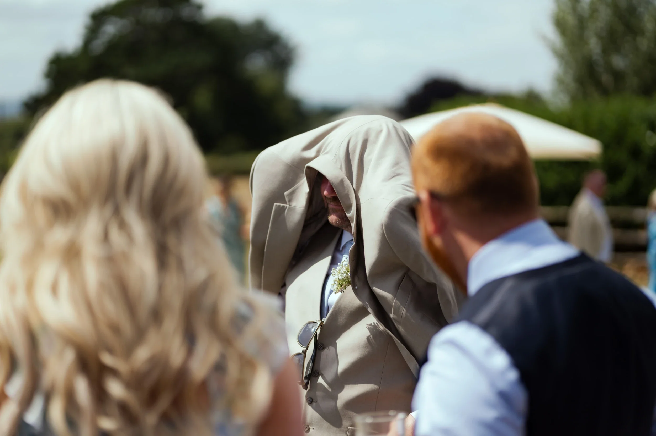 People at an outdoor event, with a man in a beige suit and a hoodie covering his face, and two other individuals, one with blonde hair and one with red hair, engaged in conversation.