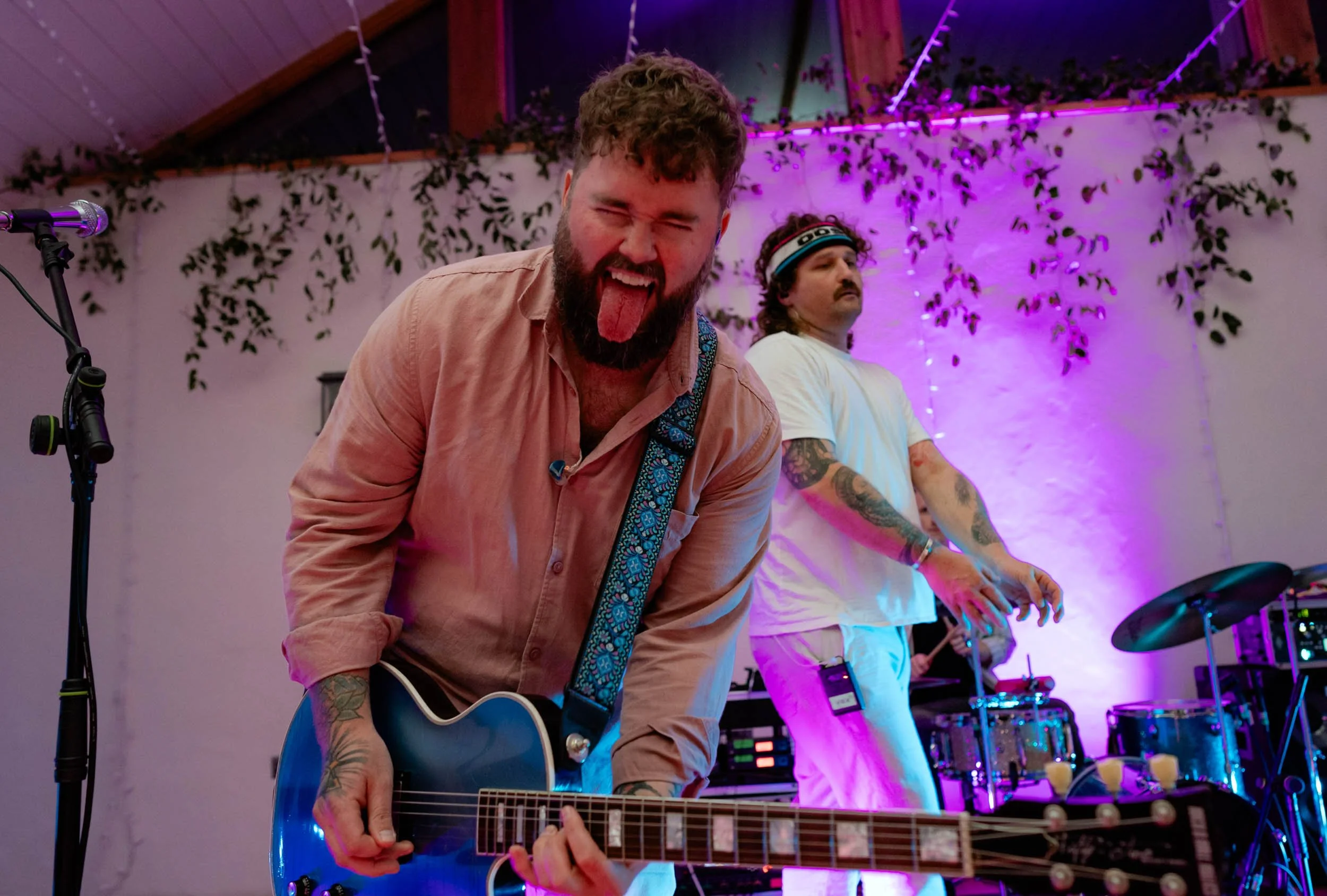 Two musicians performing on stage, one in the foreground with a blue electric guitar and sticking out his tongue, and another in the background with tattoos, wearing a white shirt and a headband, surrounded by purple and pink stage lights and musical