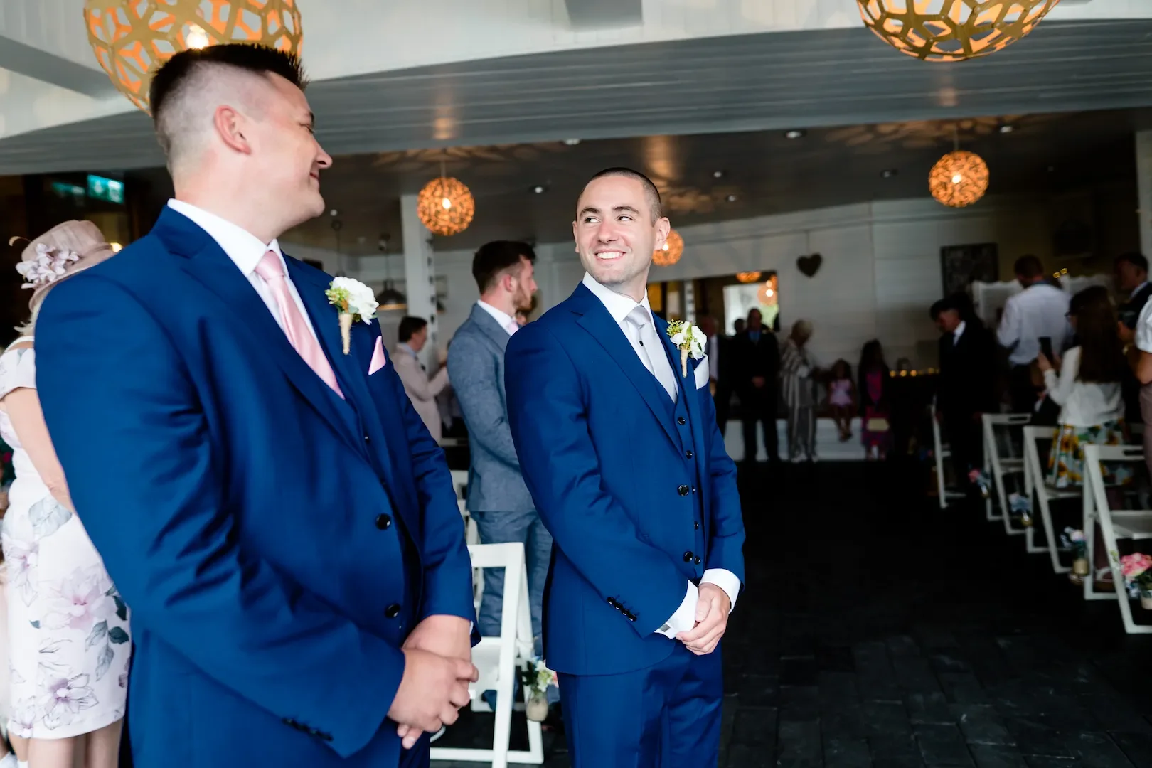 Two men in blue suits with boutonnieres on their lapels, standing inside a decorated venue, smiling at each other during what appears to be a wedding celebration.
