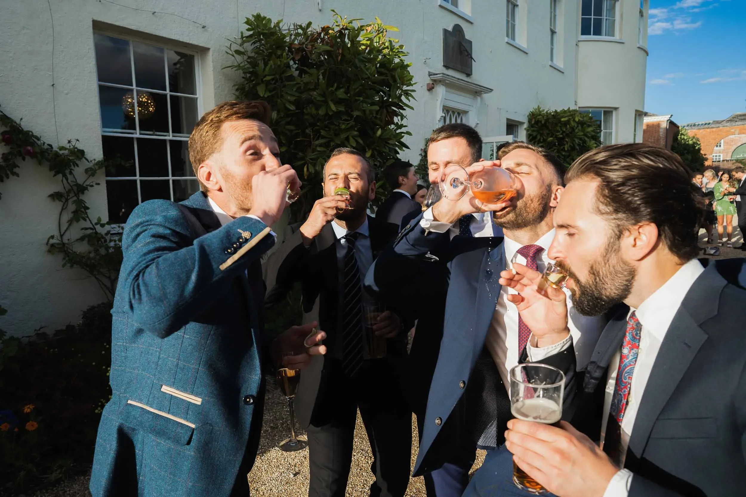 Group of men in suits at an outdoor party drinking from glasses.