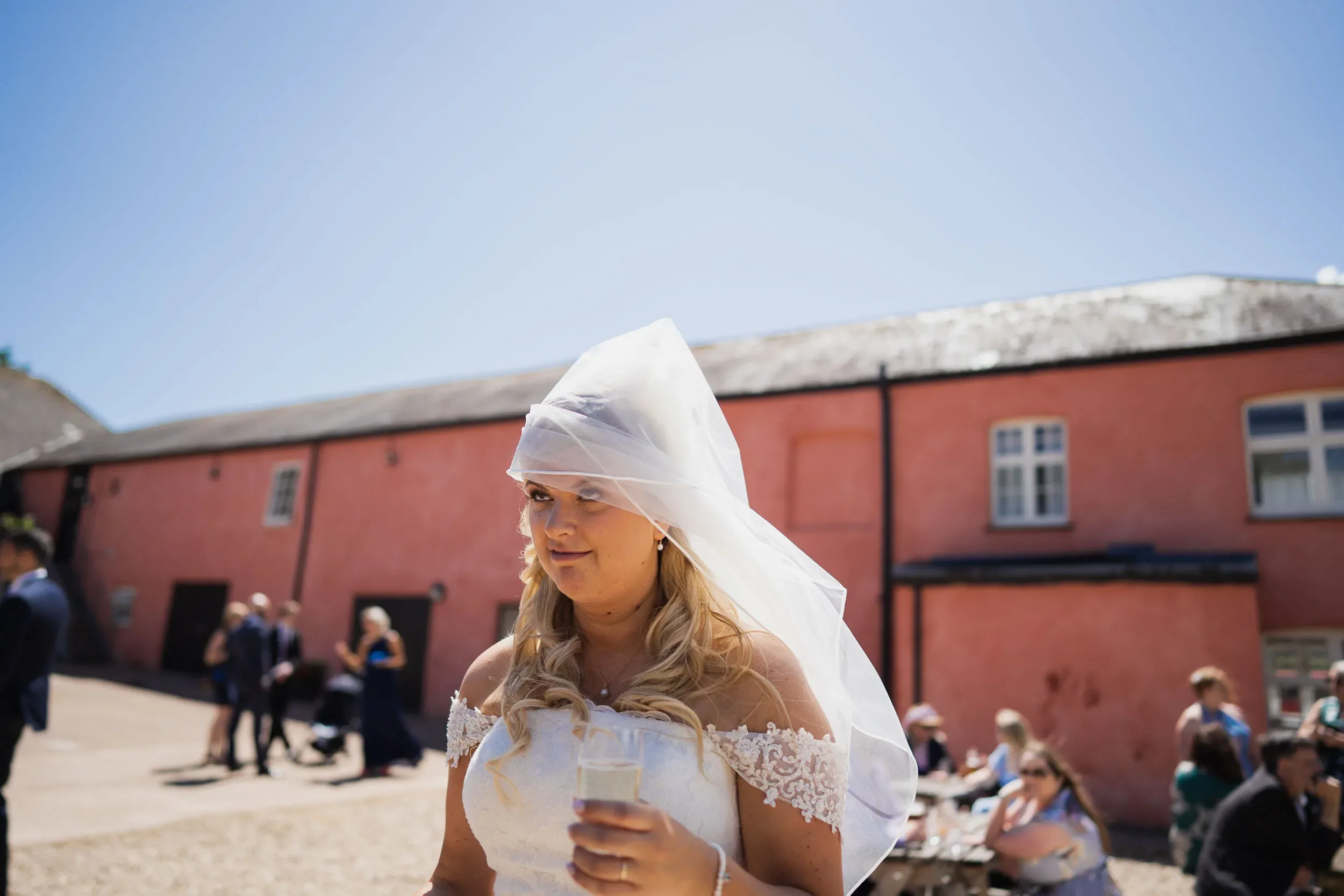 A bride wearing a white lace wedding dress and veil holding a glass of champagne at an outdoor wedding reception with guests seated at tables behind her.