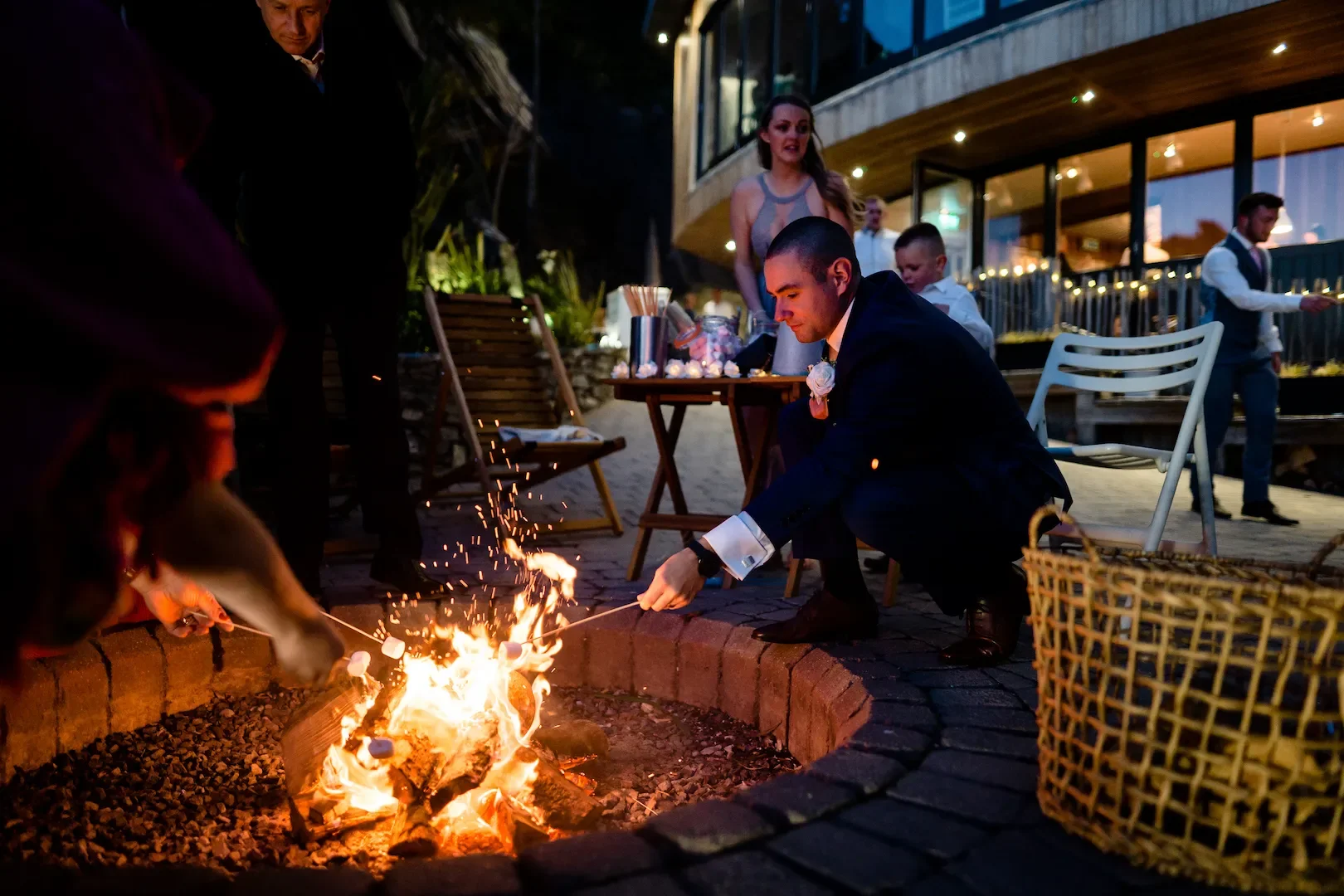 A man in a suit crouches next to a small fire pit, roasting marshmallows on long skewers during an outdoor evening event, with a woman and other guests in the background near a building with string lights.
