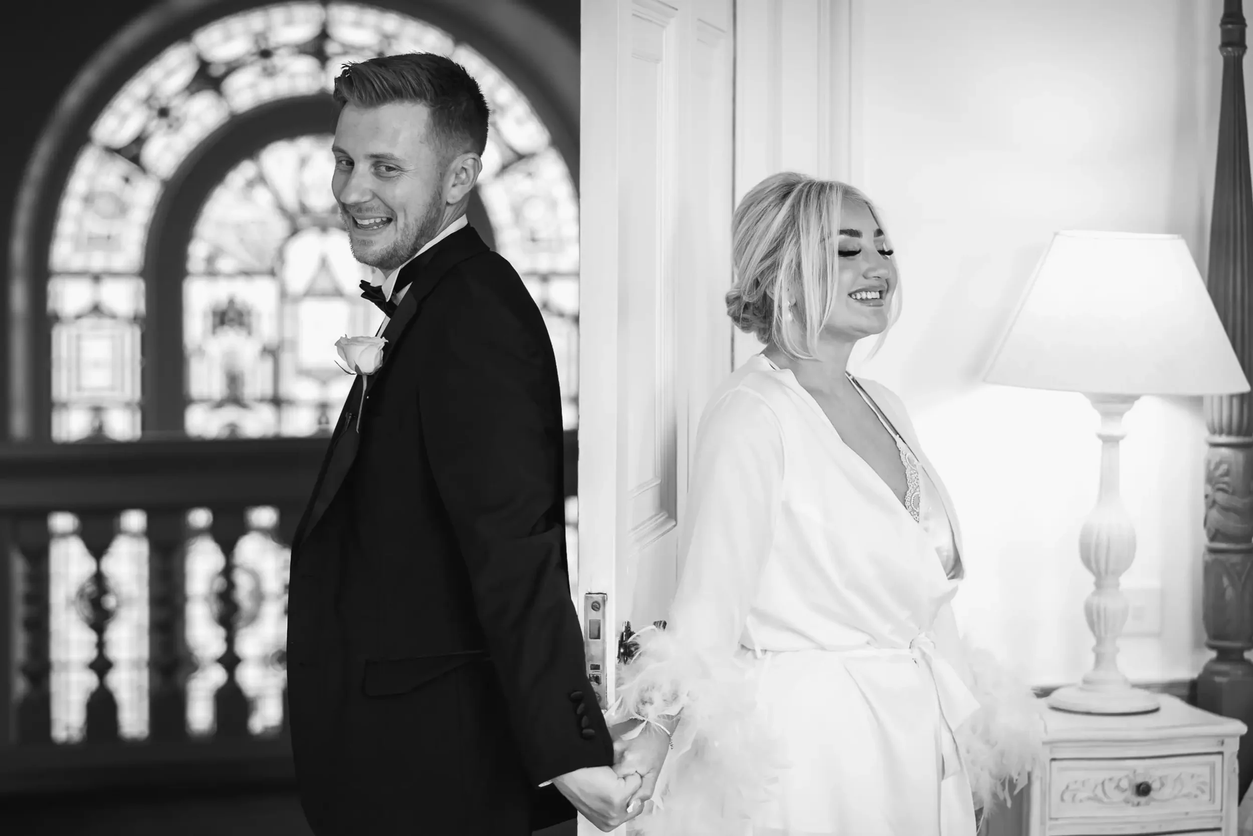 A black-and-white photo of a couple on their wedding day. The groom, dressed in a tuxedo, is smiling and holding the bride's hand behind his back. The bride, wearing a robe, is smiling with her eyes closed, leaning against a wall beside a lamp and a 