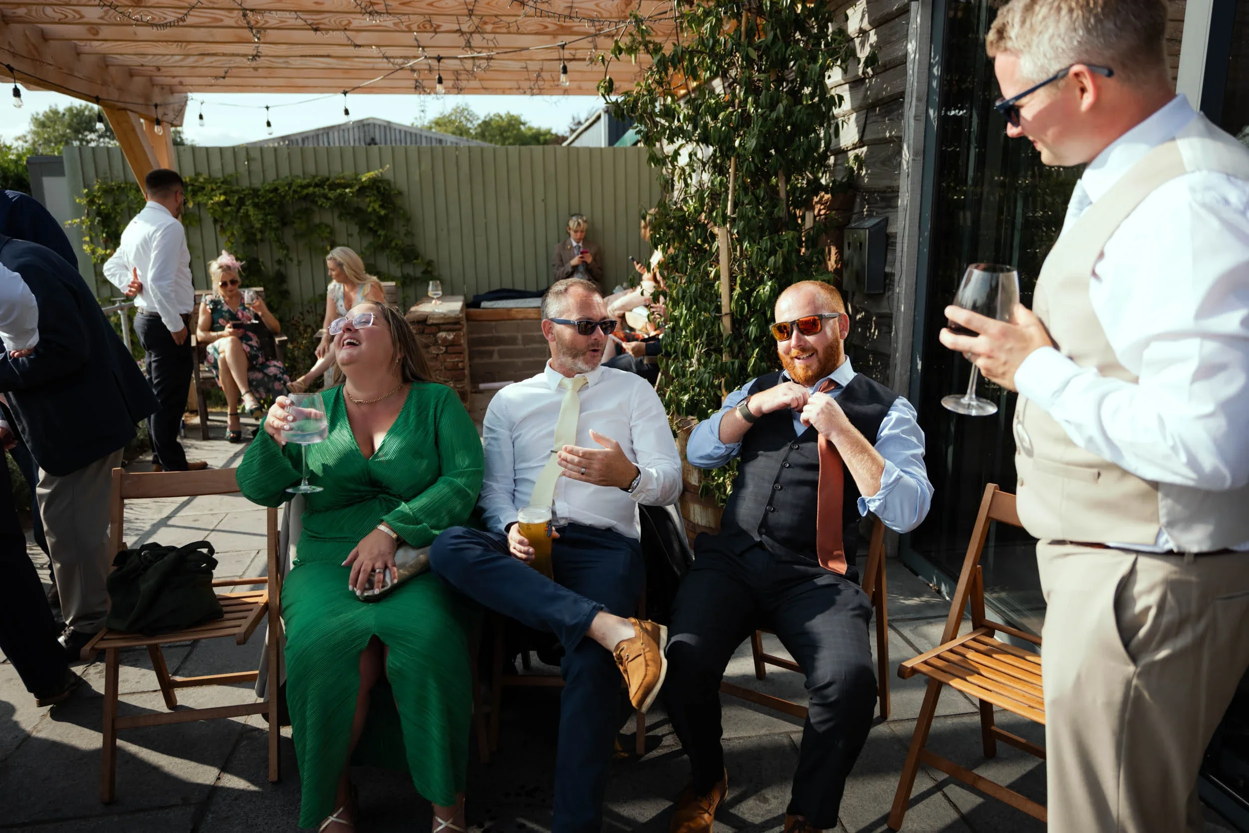 Group of people having fun at an outdoor social gathering, sitting on wooden chairs, with some standing and talking, wearing formal and semi-formal attire, on a patio with string lights overhead and a wooden pergola.