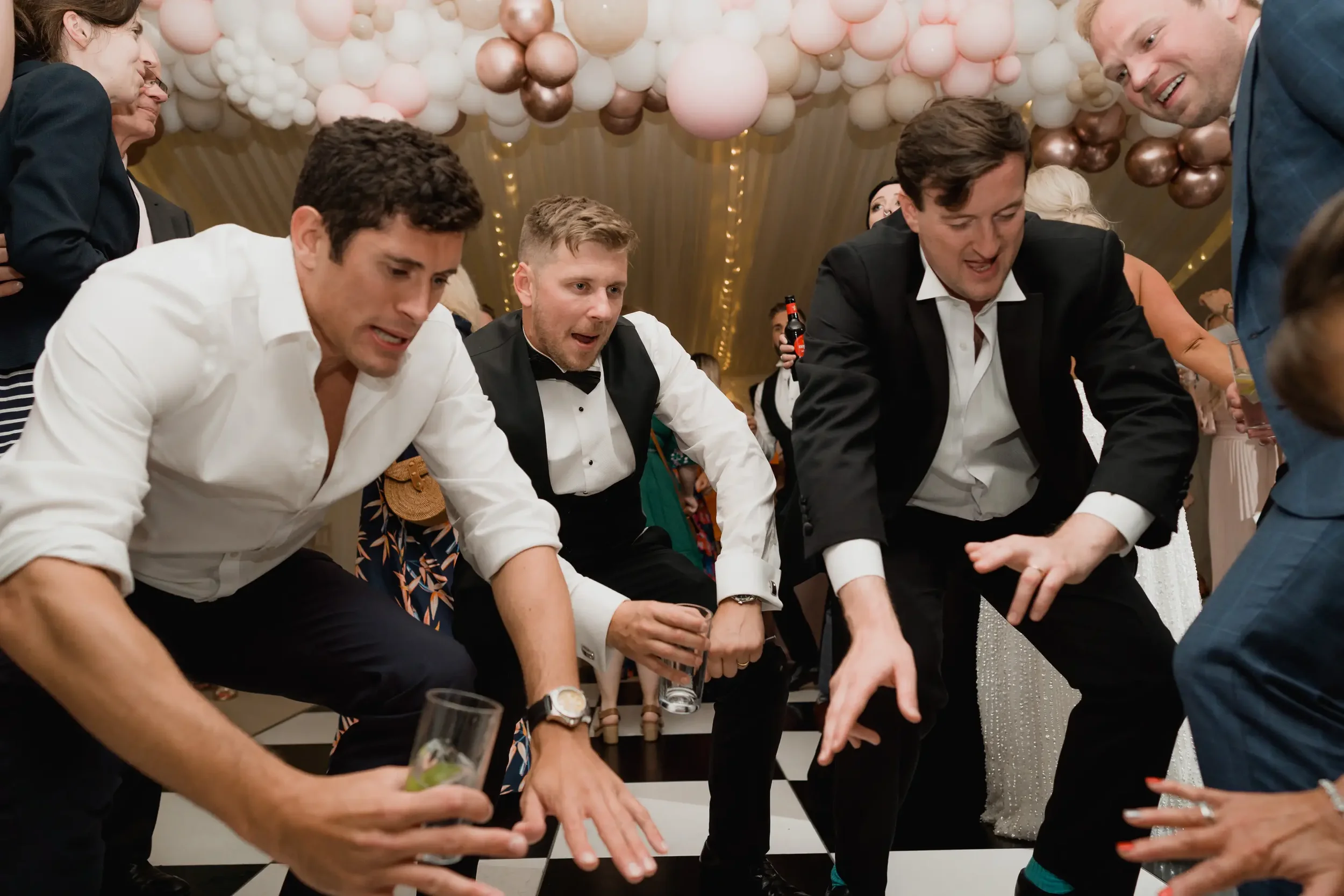 Group of men at a party dancing on a checkered floor with balloons overhead.
