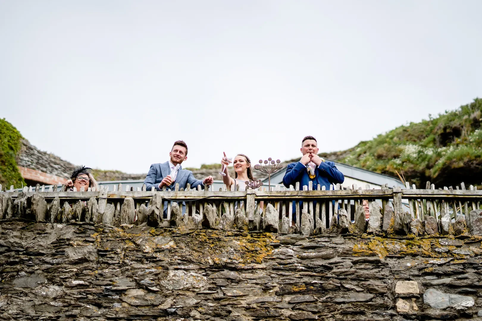 People celebrating outdoors behind a wooden fence, with a stone wall in front, on a cloudy day.