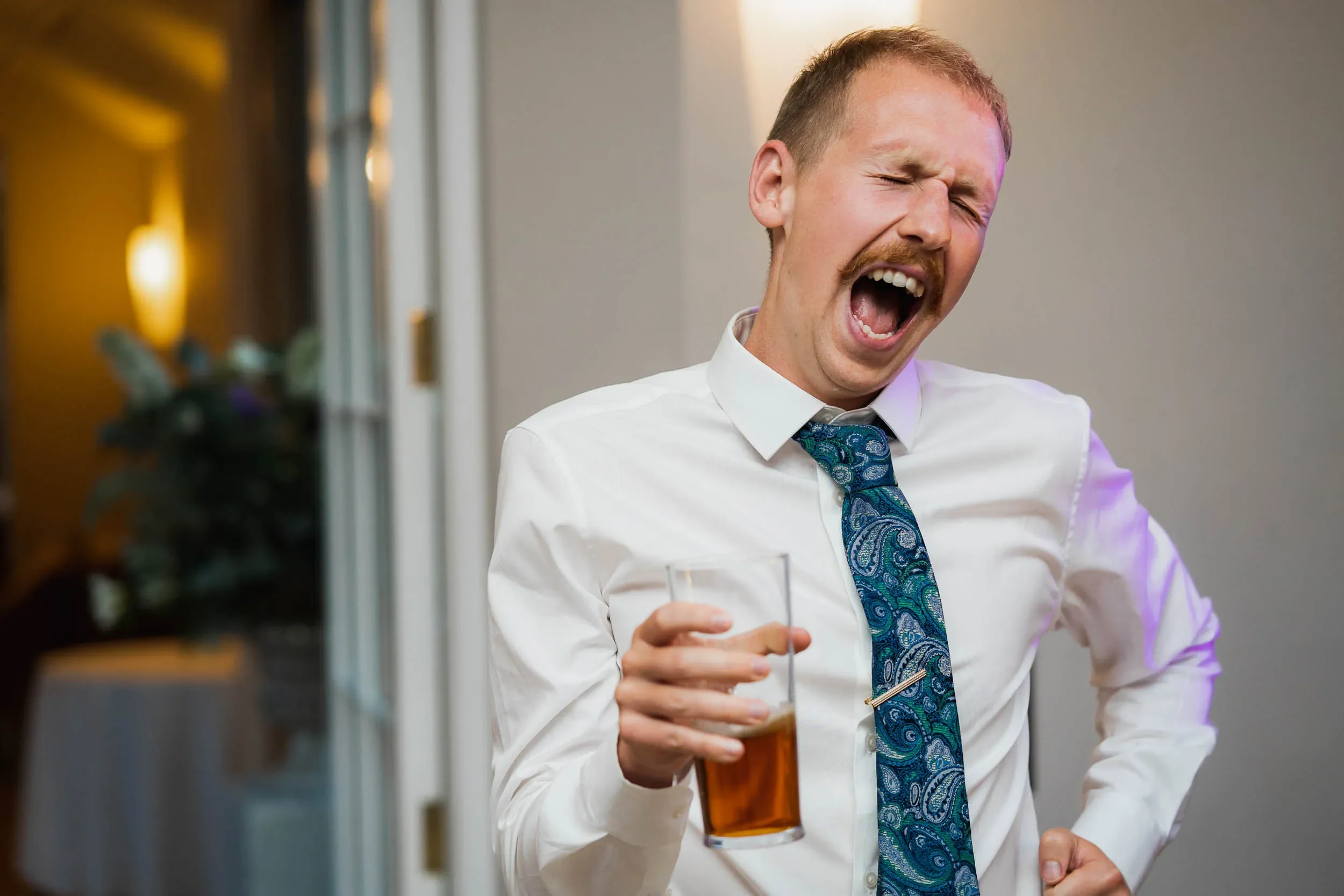 A man with a mustache wearing a white shirt and a blue patterned tie, laughing with his eyes closed and mouth wide open, holding a glass of dark beverage.