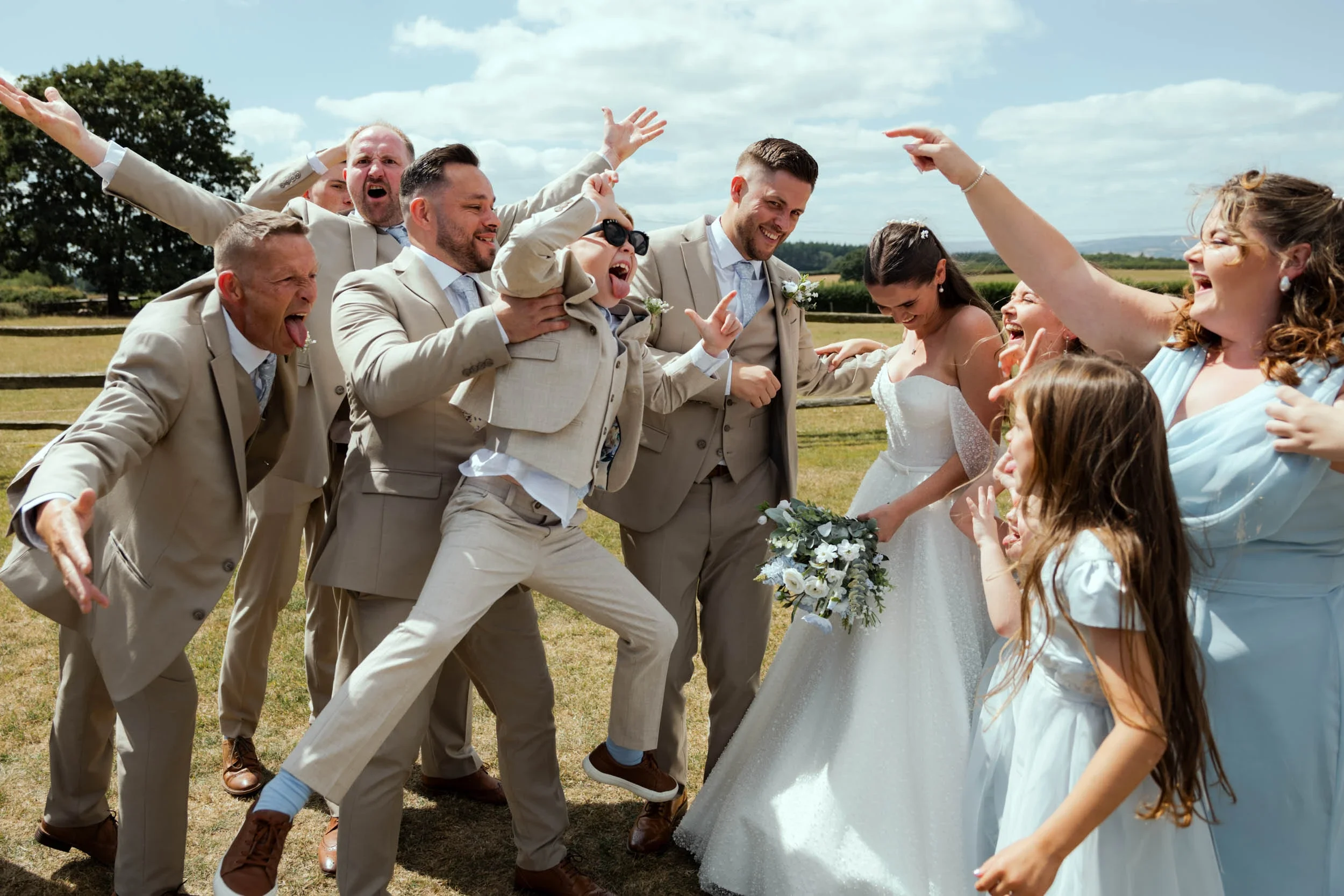 A wedding celebration outdoors with a group of people, including a bride in a white wedding dress holding a bouquet, and groom in a beige suit, surrounded by family and friends in light-colored suits and dresses, joyfully gesturing and smiling in a g