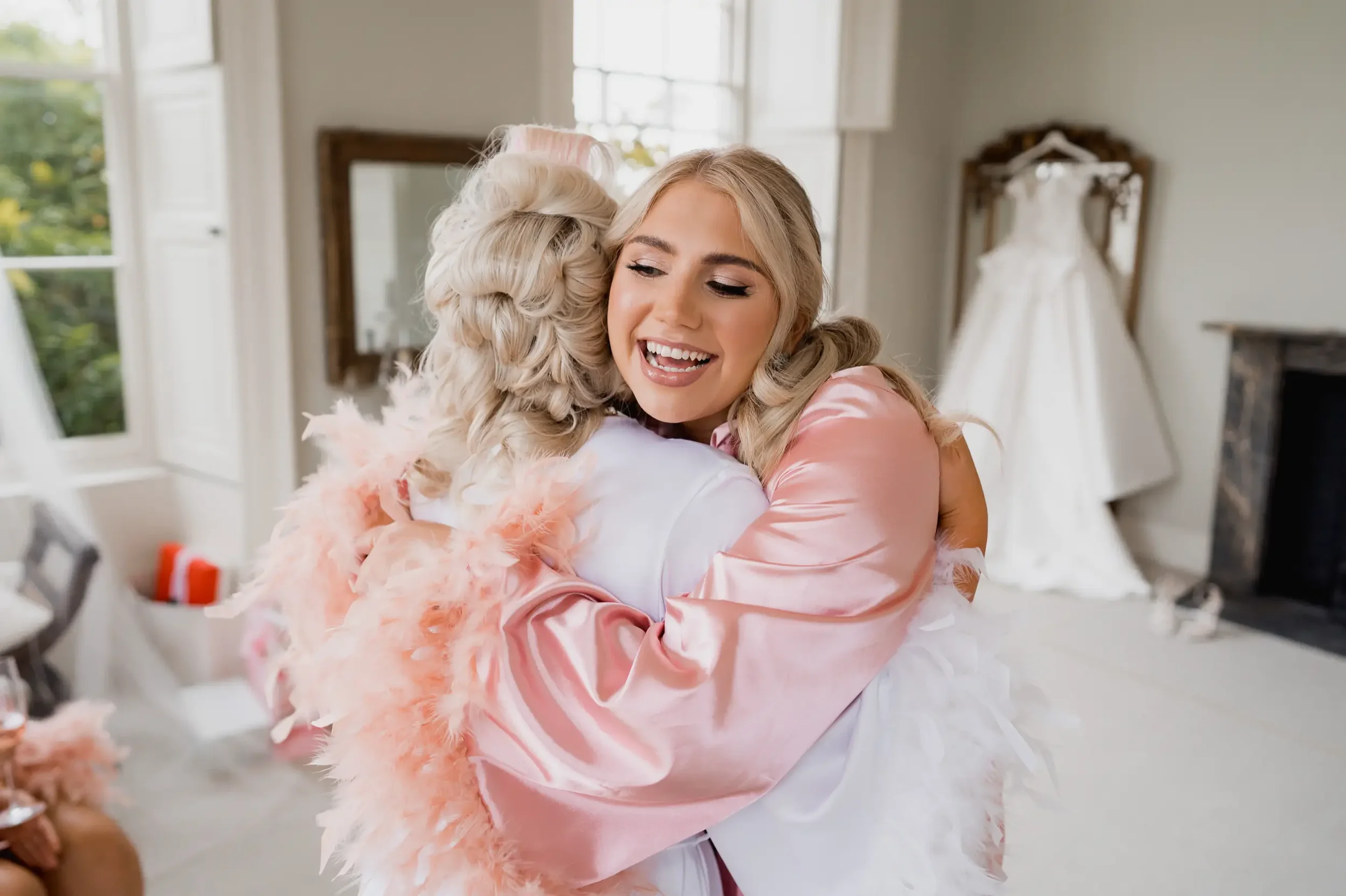 A young woman with blonde hair smiling and hugging a person dressed as a bride, who is wearing a white veil. The woman is in a pink satin robe with feather details, and the scene is set in a bright room with a wedding dress hanging in the background.
