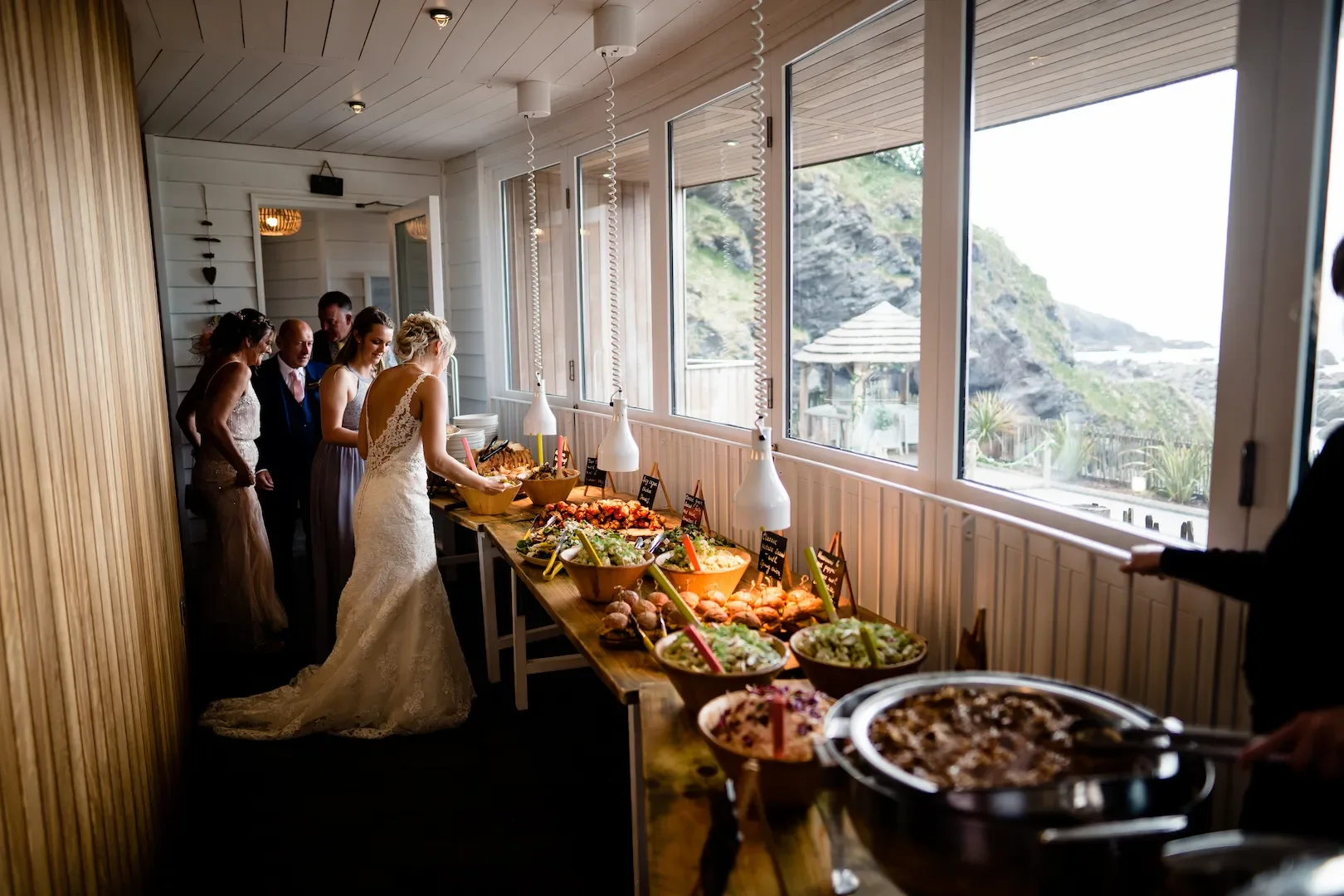 Wedding reception with bride and guests at a buffet table with salads and other dishes, overlooking a scenic outdoor view.