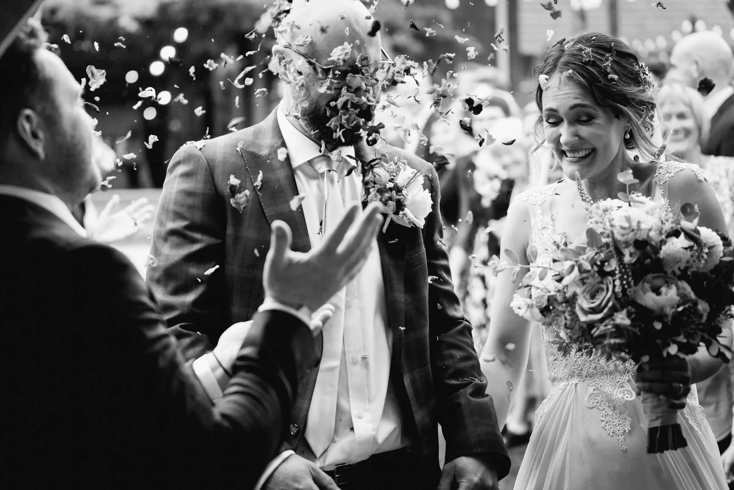 Black and white photo of a wedding celebration with a bride holding a bouquet of flowers and a groom, both smiling, while guests throw flower petals.