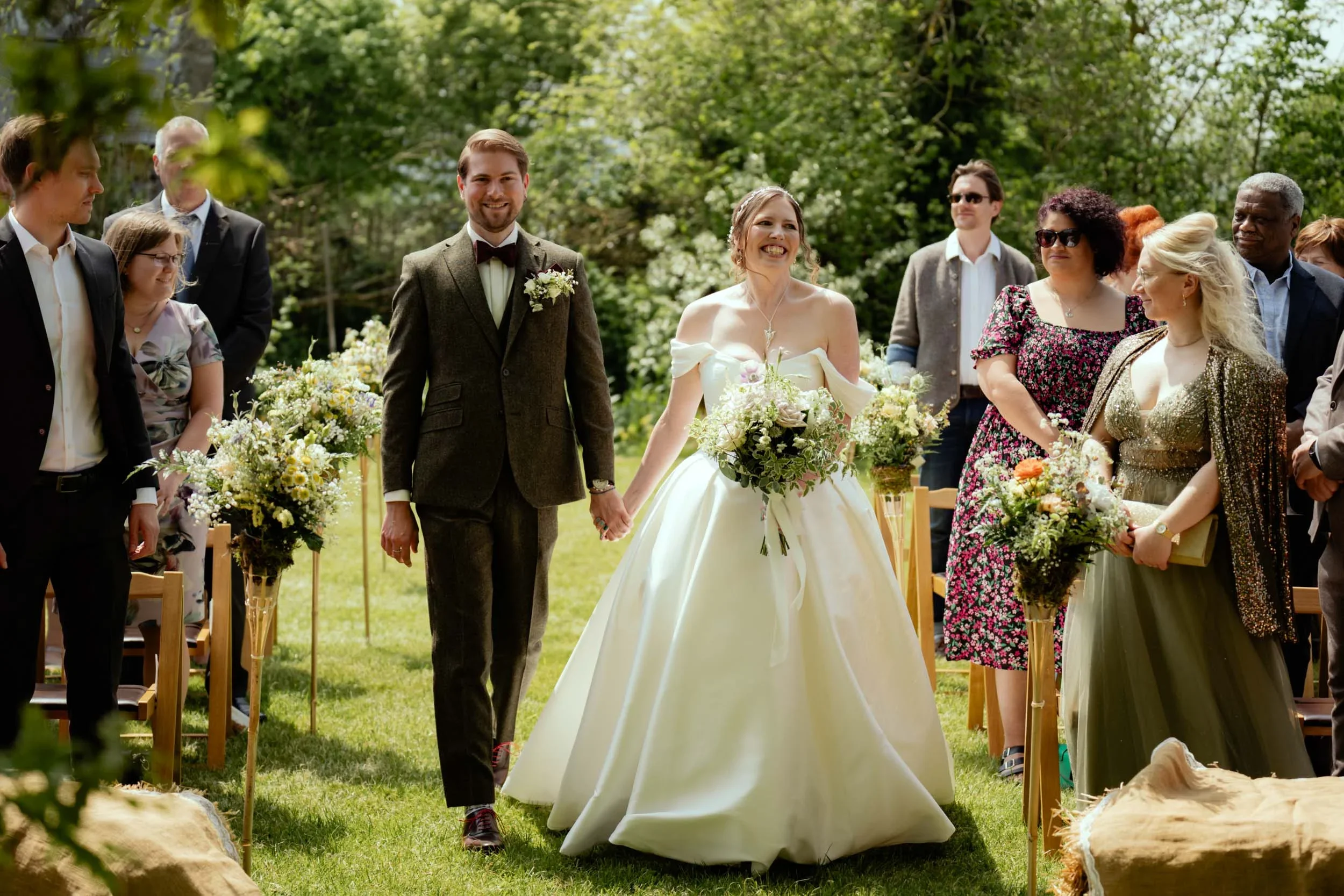 Bride and groom holding hands walking down the aisle during a wedding ceremony outdoors with guests seated on either side, greenery and trees in the background.