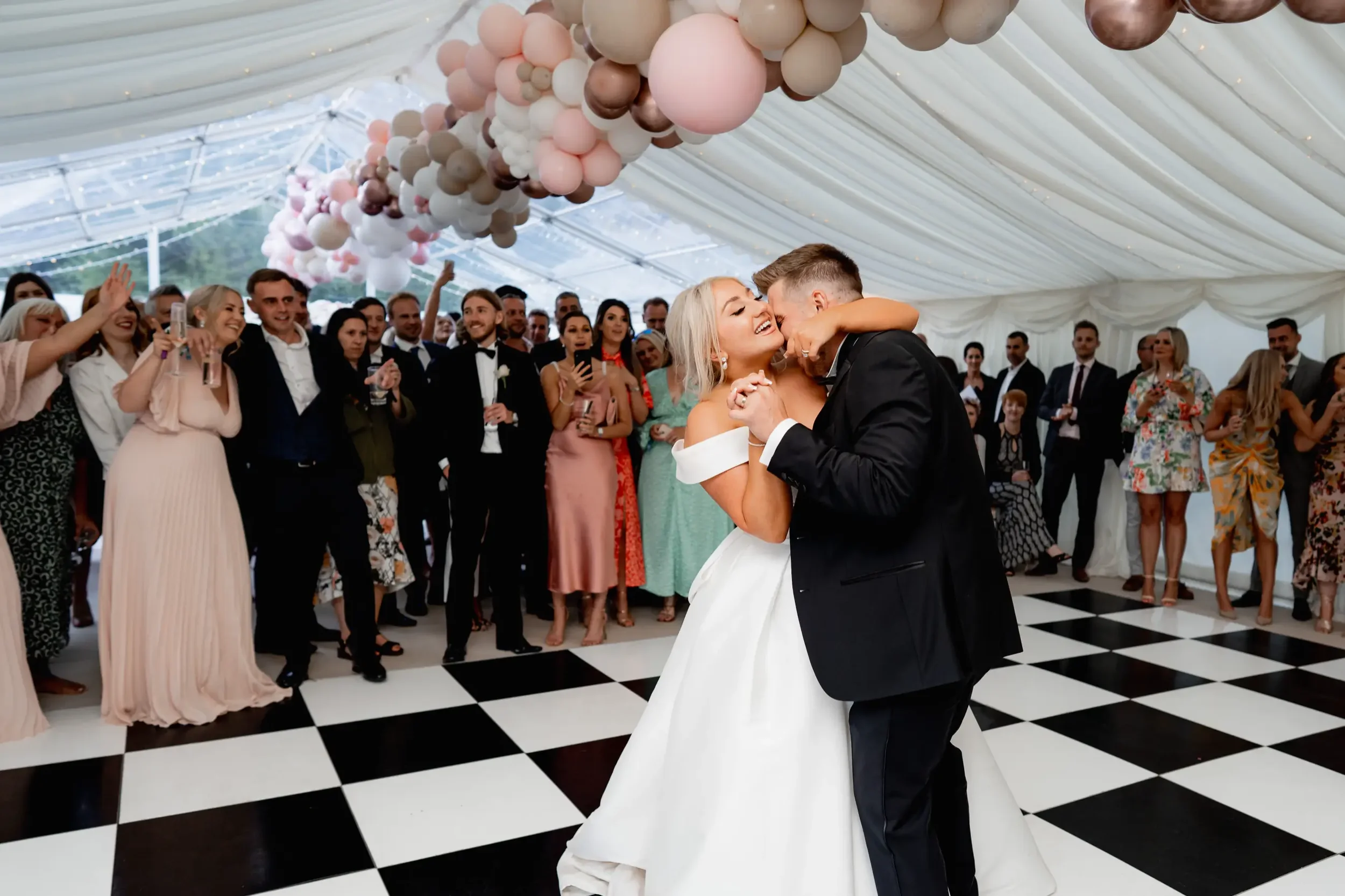 A newlywed couple shares a dance at their wedding reception, surrounded by family and friends in a decorated tent with balloon accents.
