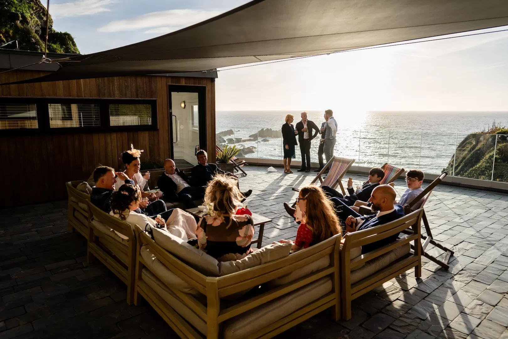 People gathered on a rooftop patio with ocean views, some sitting on outdoor furniture and others standing near the railing, enjoying a social event during sunset.