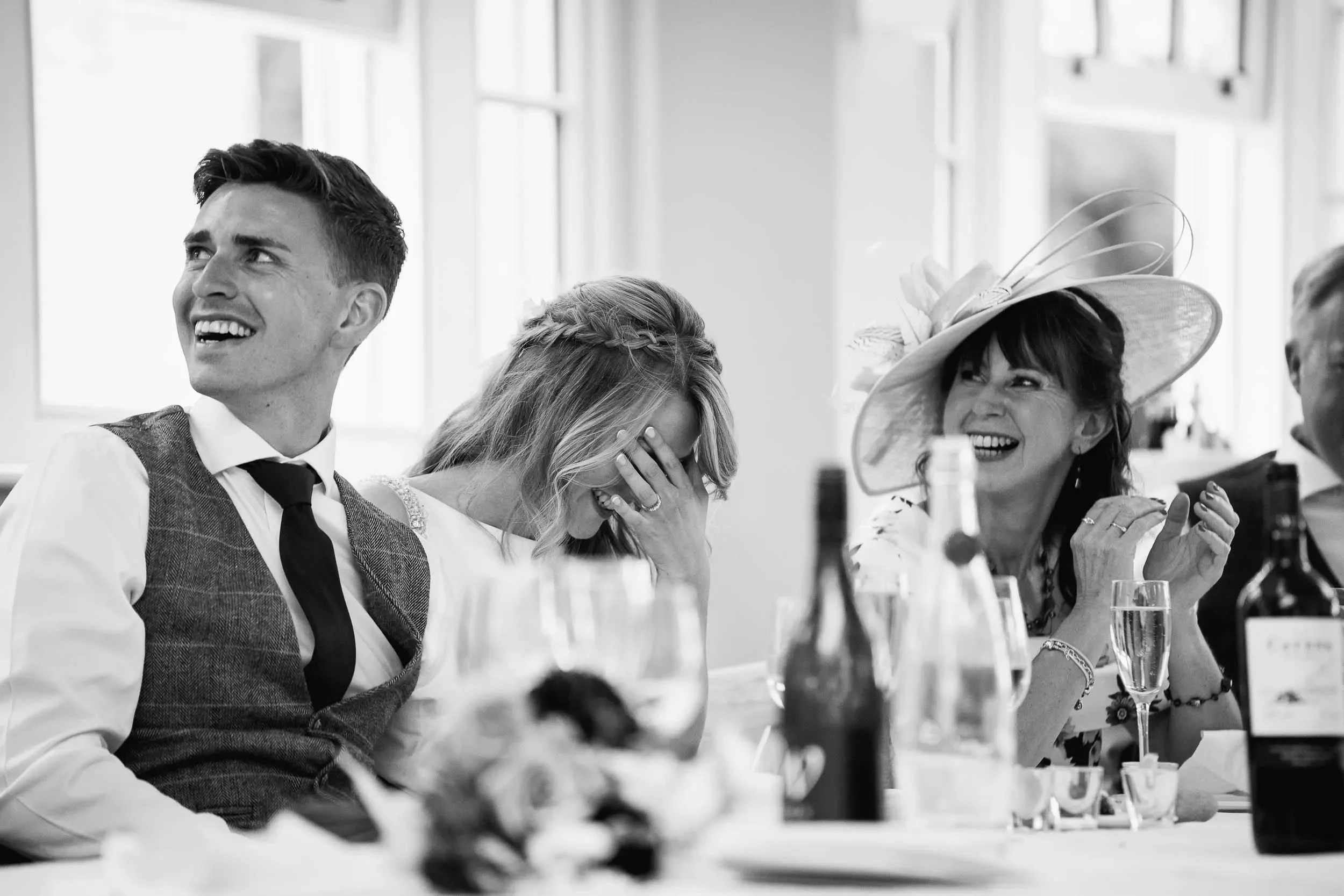 People at a celebration, with two women laughing and a man smiling, sitting at a table with drinks and bottles, one woman wearing a large hat, in a bright room with windows.
