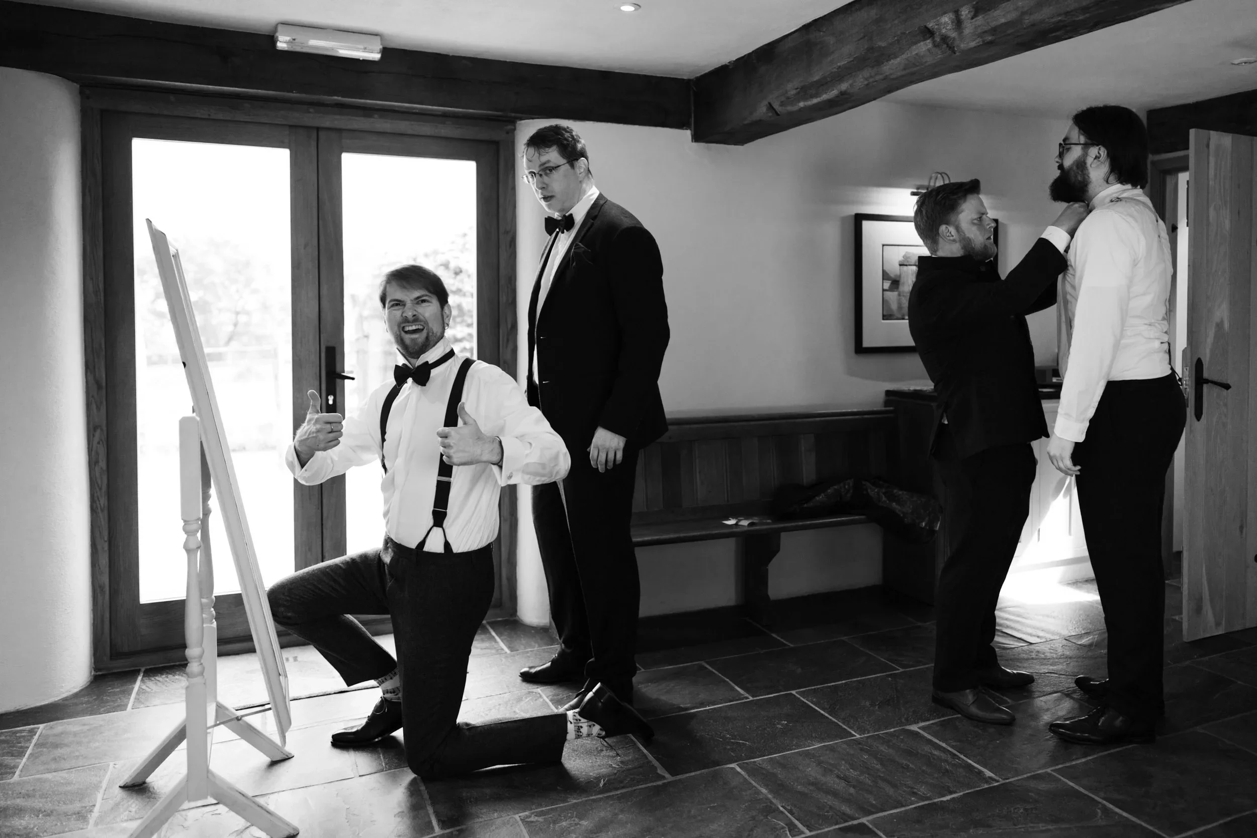 Four men in formal attire inside a room, with one kneeling and three standing. Two are adjusting each other's ties, one is giving a thumbs-up, and another is observing. The room has a wooden floor, a window with glass doors, and a bench along the wal