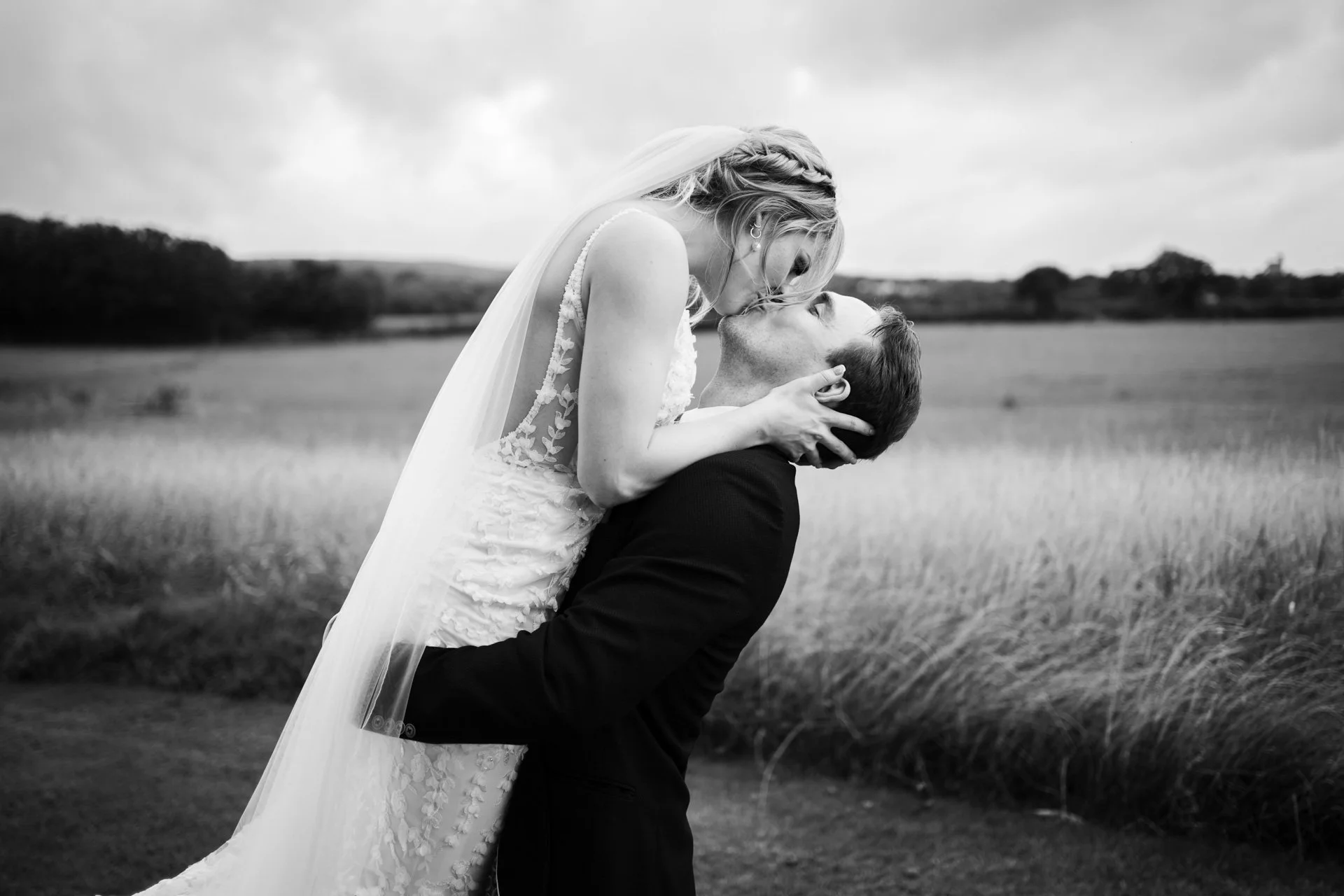 A bride and groom sharing a kiss outdoors, with the groom holding the bride up. The bride is wearing a lace wedding dress and veil, and the groom is dressed in a dark suit. They are in a field with tall grass and trees in the background.