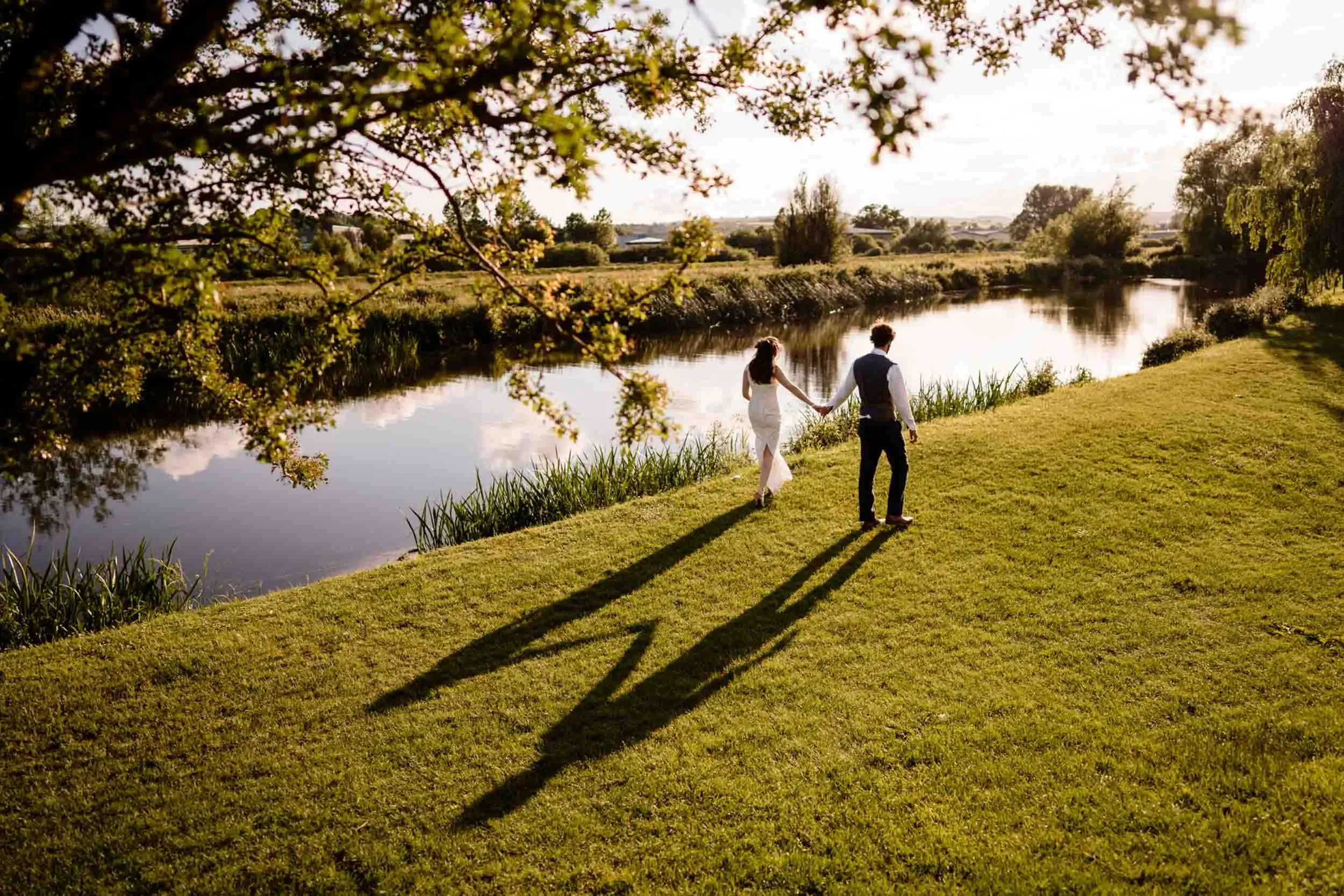 A couple walking hand in hand along a grassy bank by a river at sunset, with their long shadows cast on the ground.