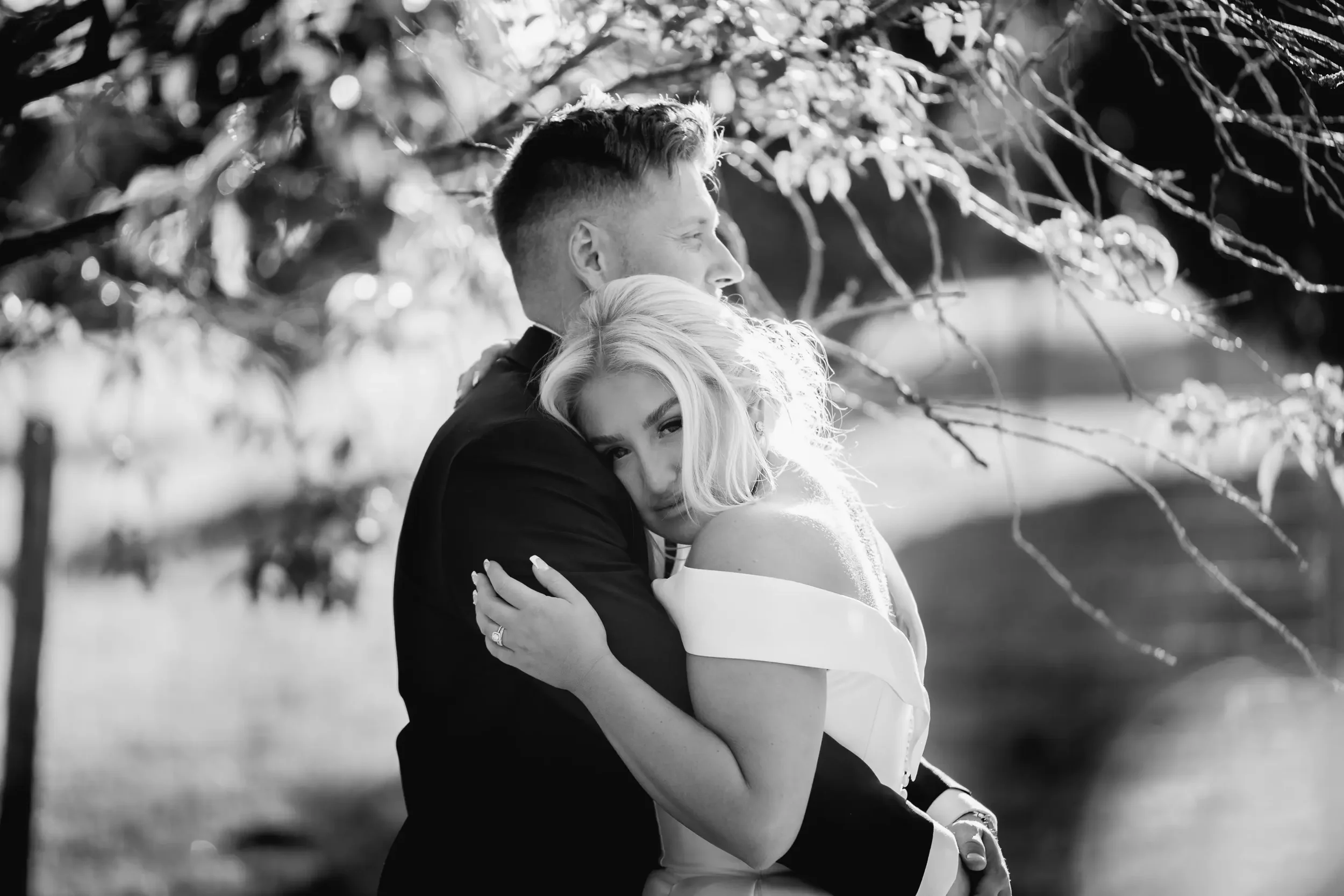 A black and white photo of a couple hugging outdoors beneath a tree, with the woman resting her head on the man's shoulder.