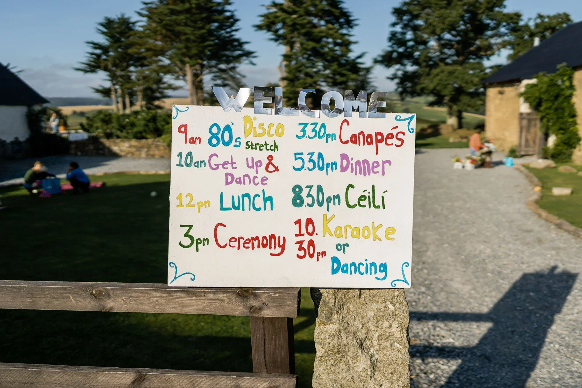 A colorful outdoor event schedule signboard listing activities from 9 am to 10:30 pm, including disco, breakfast, get up & dance, lunch, ceremony, canapés, dinner, céilí, karaoke, and dancing, set against a rural landscape with trees, stone buildings
