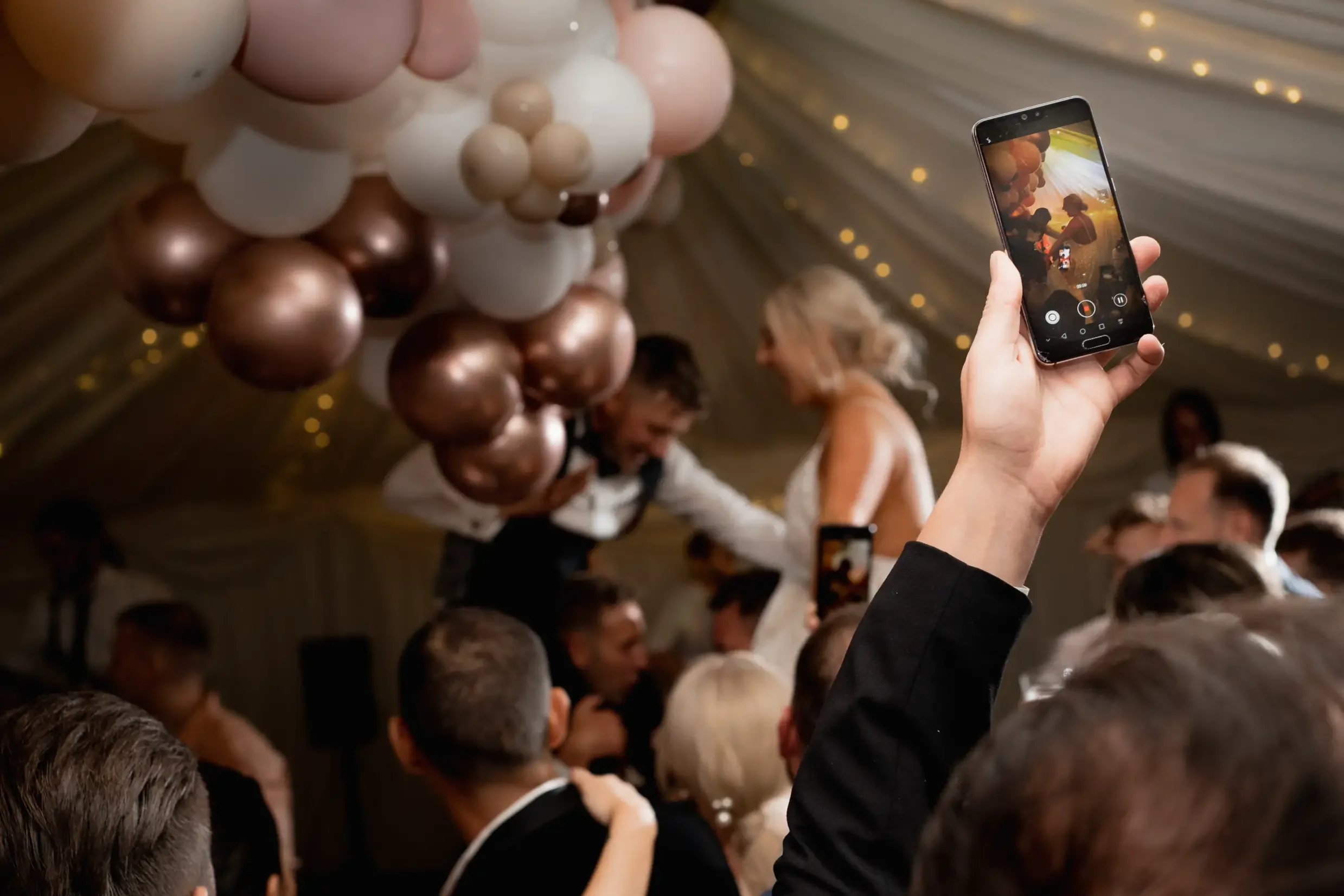 Person taking a photo of a wedding couple on a smartphone at a celebration with balloons and decorative lighting.