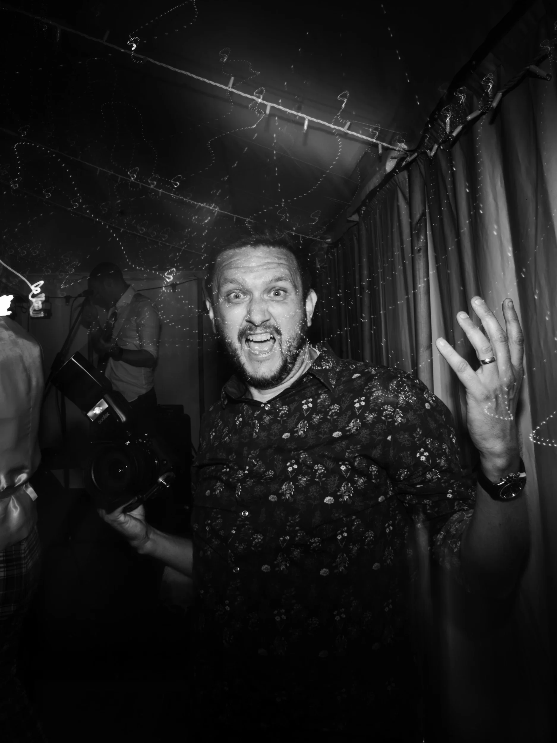 A man with a beard and mustache, wearing a floral shirt, is making a wild facial expression at a party or event, holding a camera in his right hand, with his left hand raised showing his fingers, in a dimly lit room with string lights and curtains.