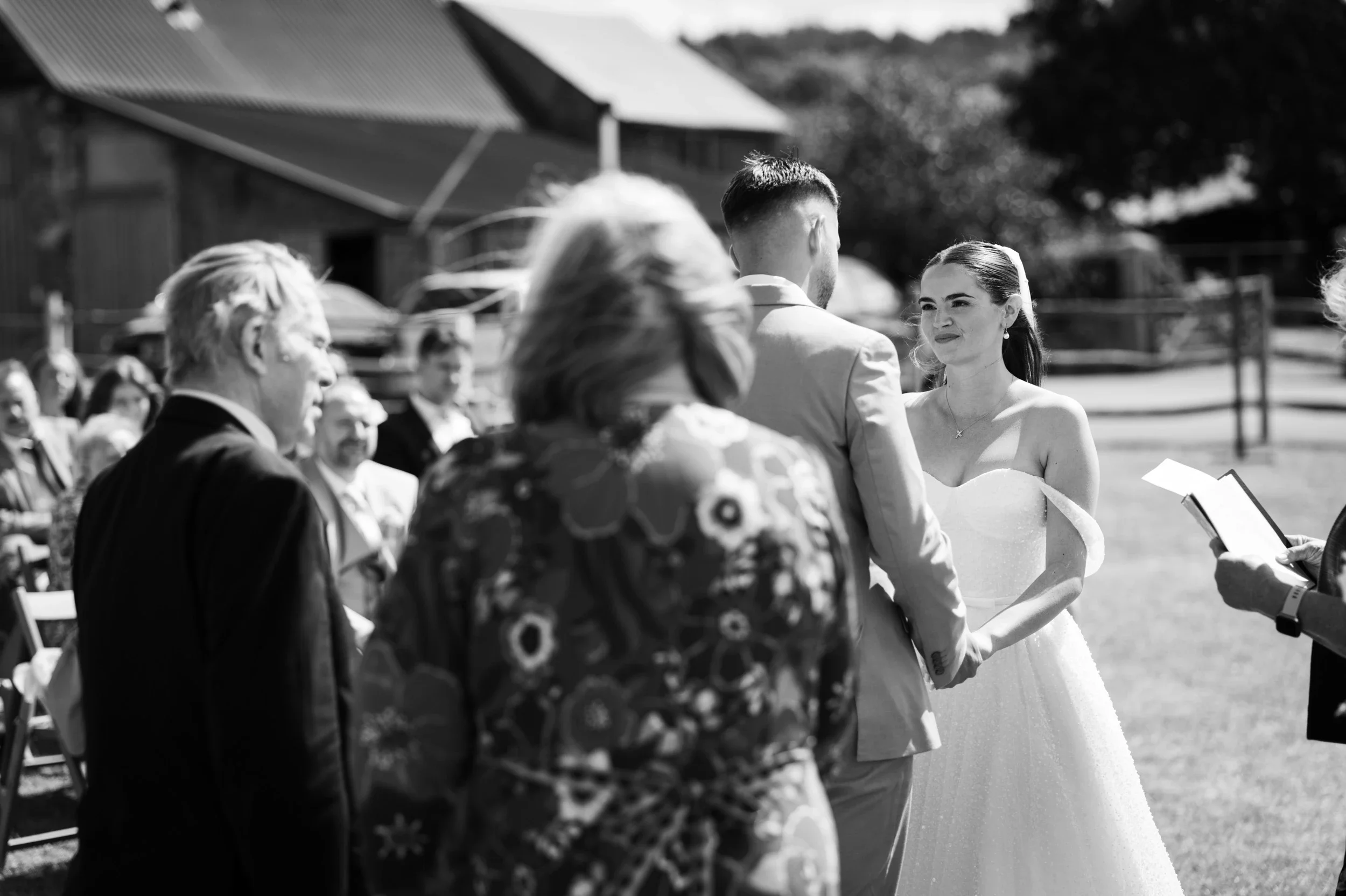A black-and-white photo of a wedding ceremony outdoors, with a bride and groom exchanging vows, surrounded by guests seated on chairs.