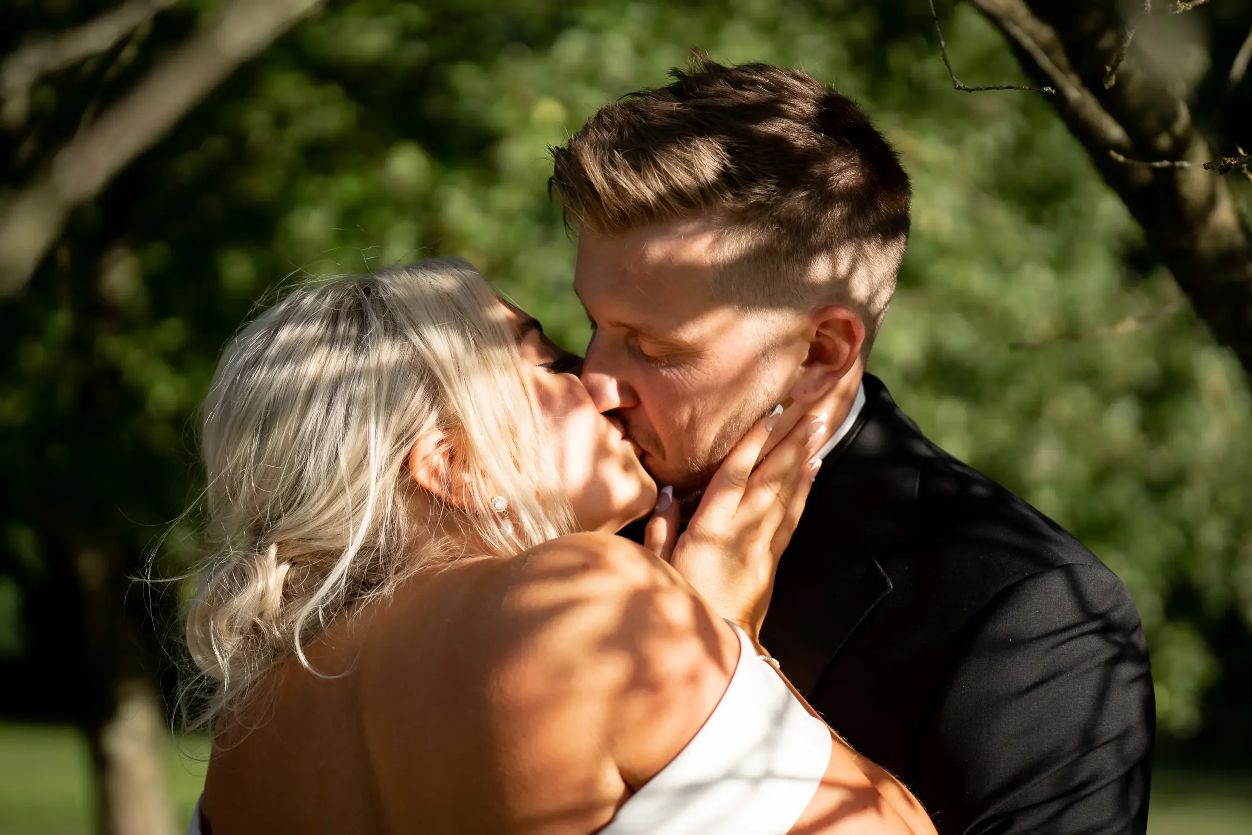 A couple kissing outdoors with greenery in the background, the woman has blonde hair and is wearing earrings, and the man has short brown hair, wearing a dark jacket.