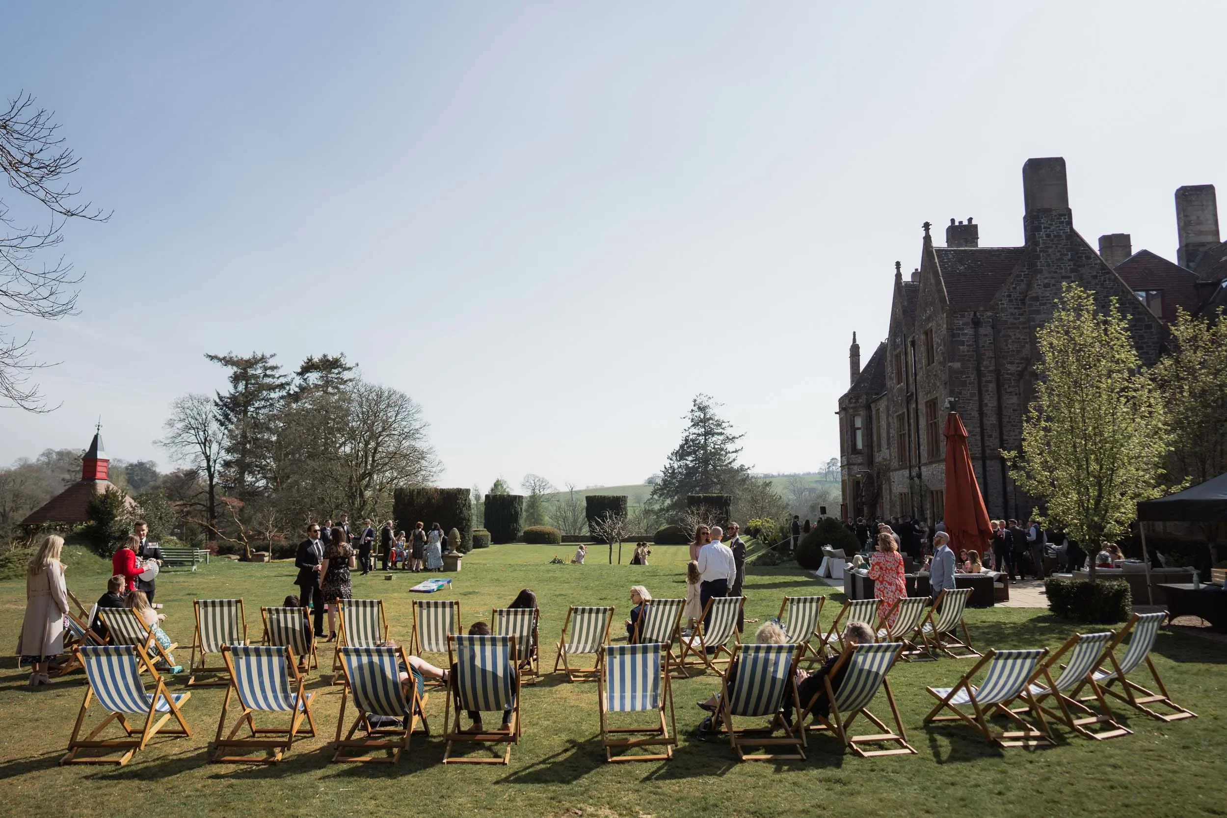 Outdoor gathering at huntsham court with people dressed formally on a lawn with striped deck chairs, in front of a large stone building, with trees and a clear sky.