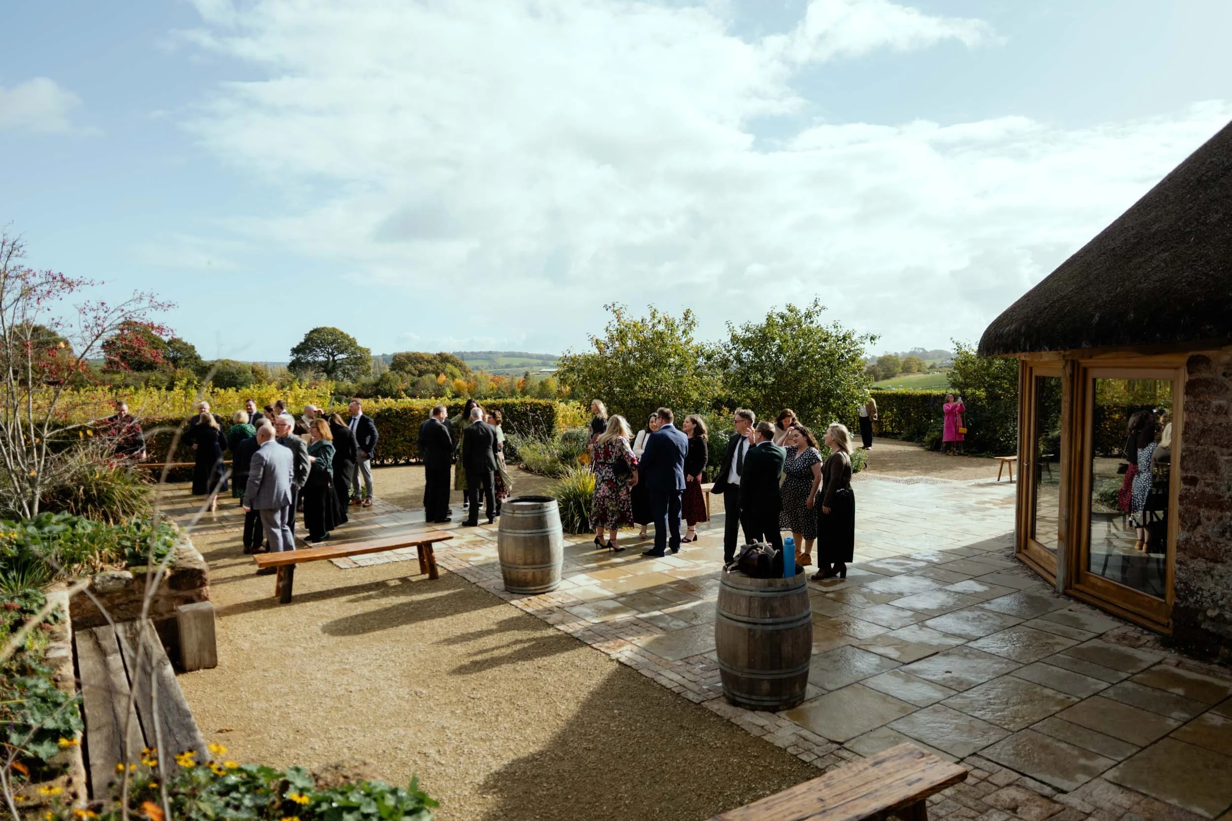 People gathered outdoors for a social event on a sunny day, with a stone building with a thatched roof and scenic landscape in the background.