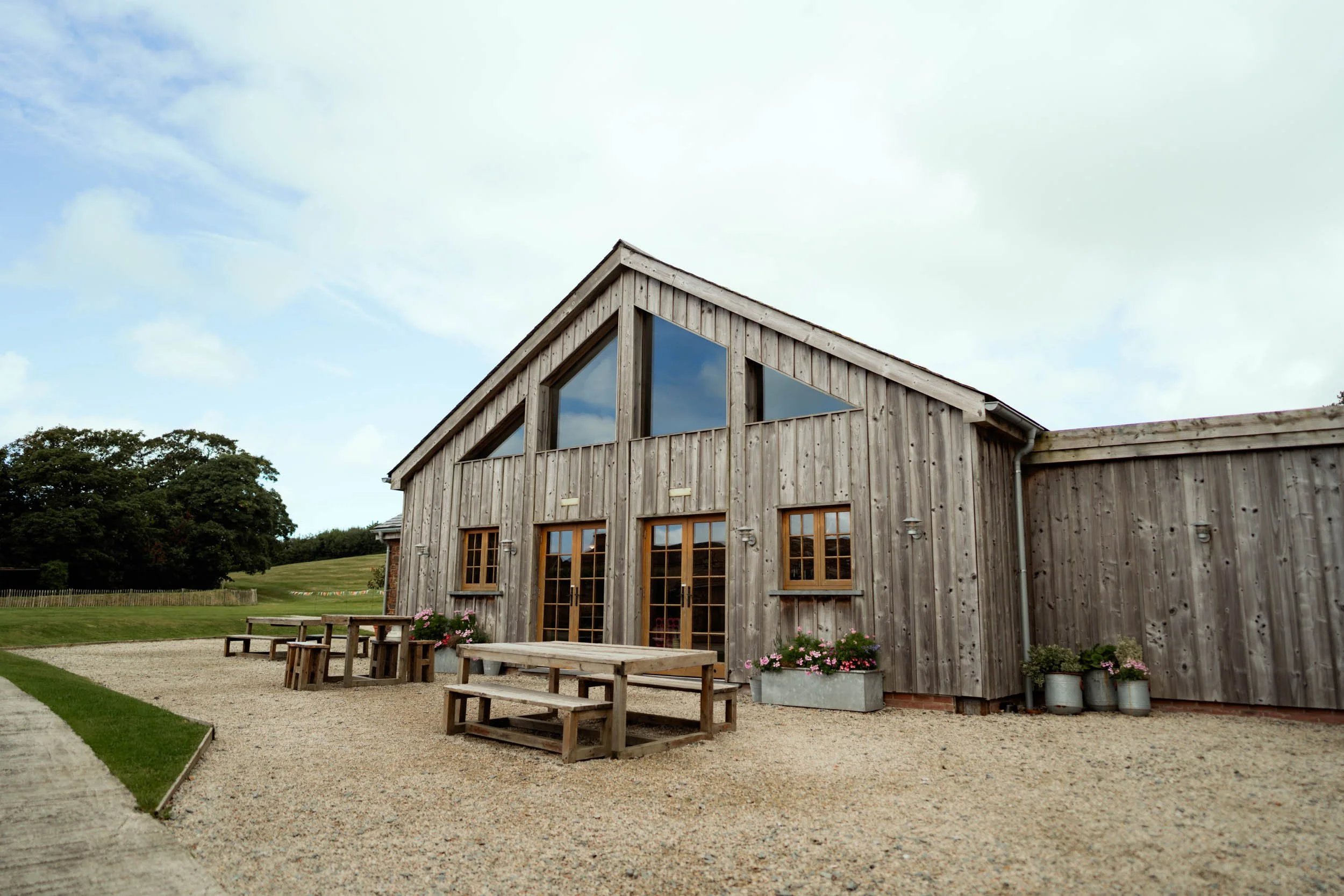 A rustic wooden building with large triangular and rectangular windows, outdoor seating with benches and a table, and flower pots, set in a grassy area with a gravel patio and a cloudy sky.
