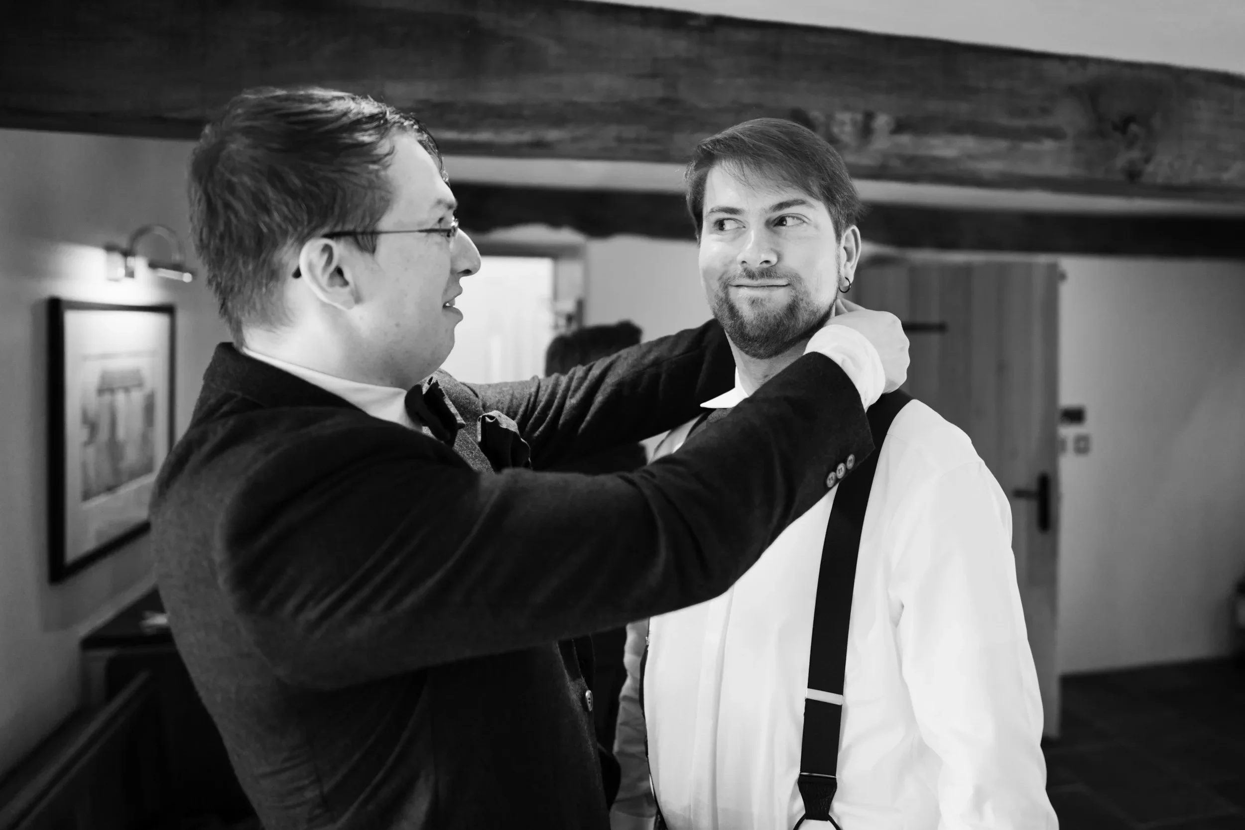 Two men in formal attire—one adjusting the other's collar—inside a room with a framed picture on the wall.