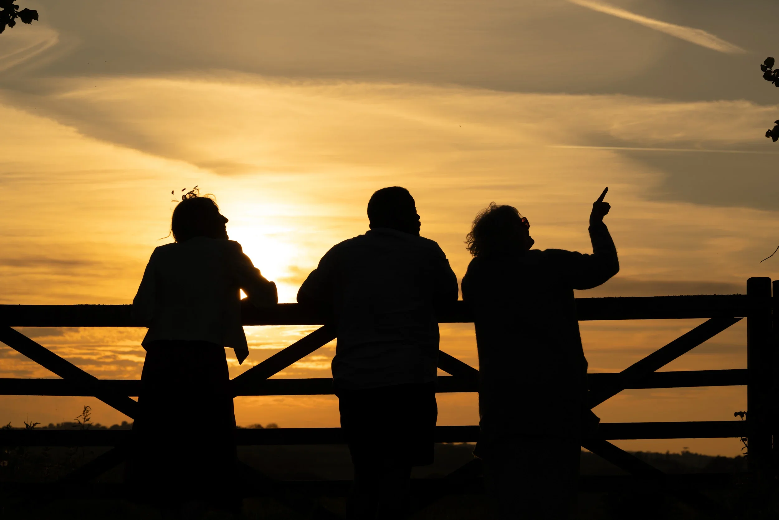 Silhouettes of three people standing by a fence during sunset, with one person gesturing upward.