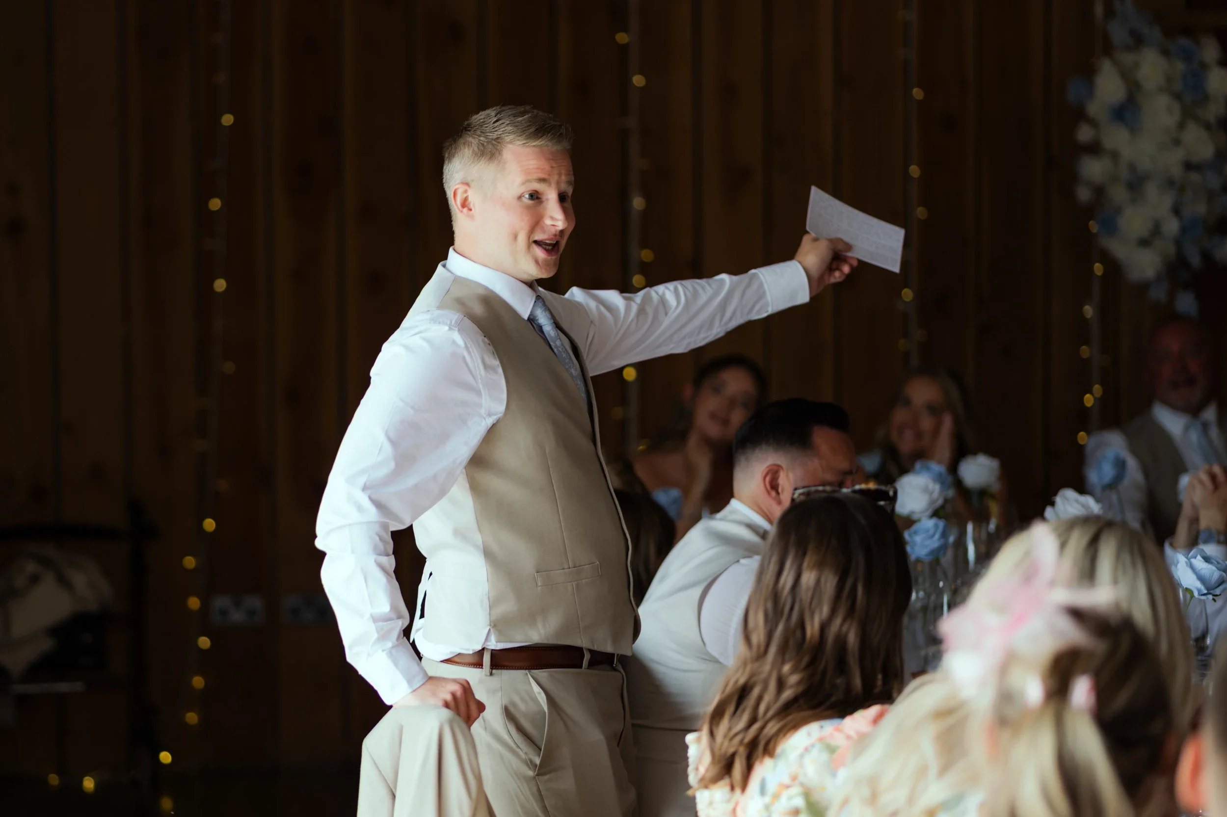 A young man in a beige vest and tie stands and talks at a wedding reception, holding a piece of paper while guests seated around him listen.