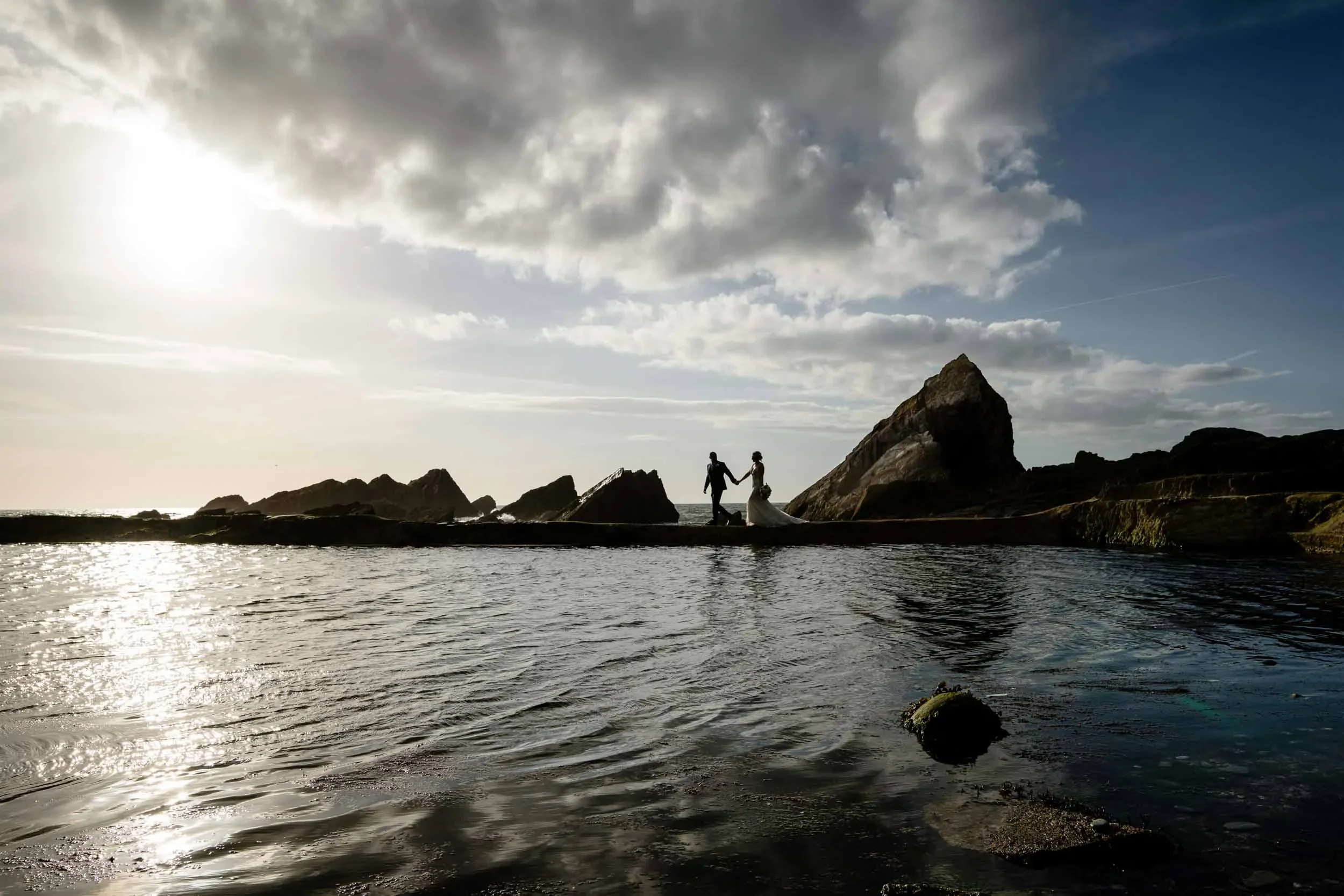 A silhouette of a couple holding hands and walking along a rocky shoreline at sunset, with the sea in the foreground and partly cloudy sky overhead.