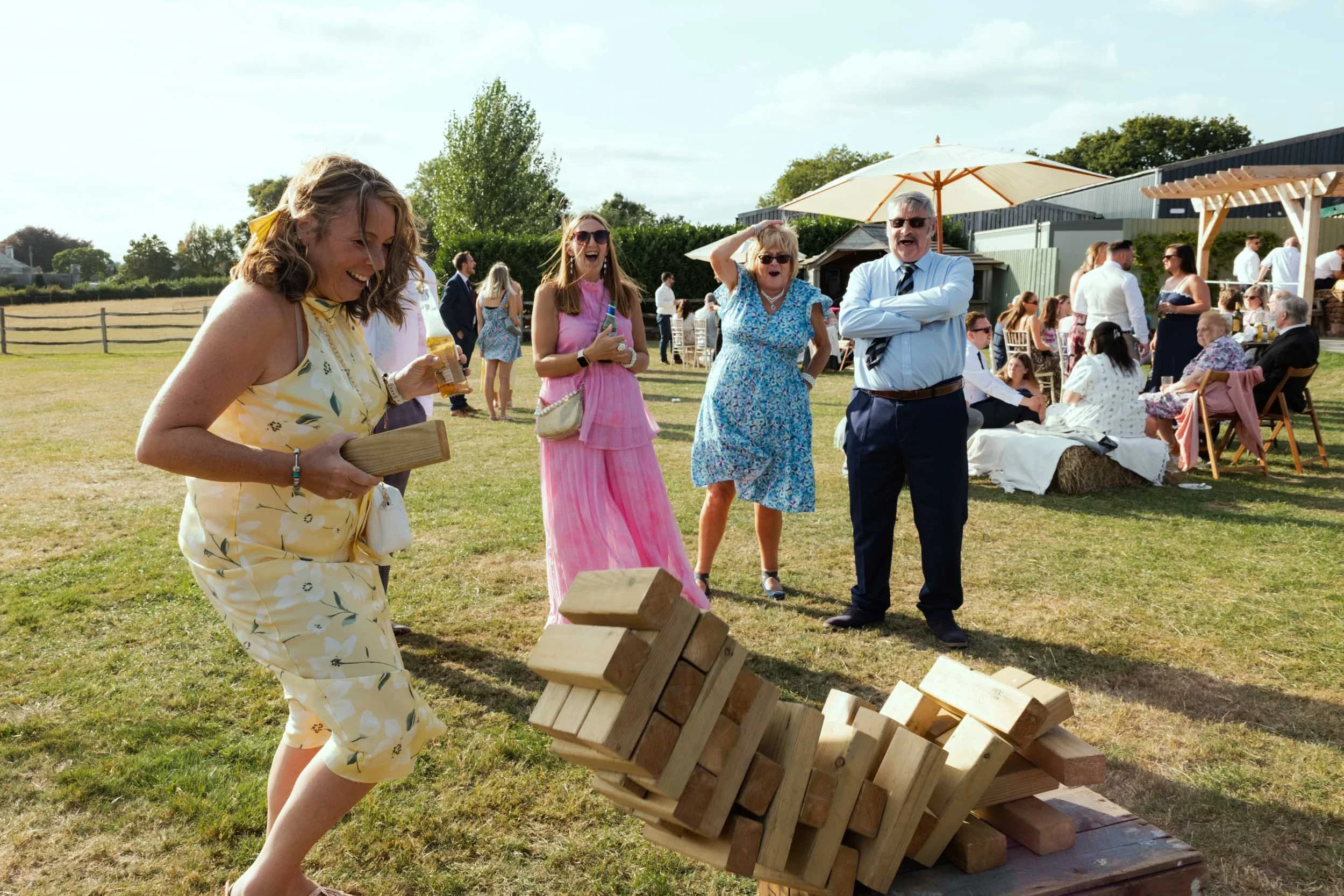 A woman in a yellow floral dress is playing giant Jenga at an outdoor party, with a group of guests watching and laughing. The event is taking place on a grassy field with tables, chairs, and people socializing in the background under umbrellas.