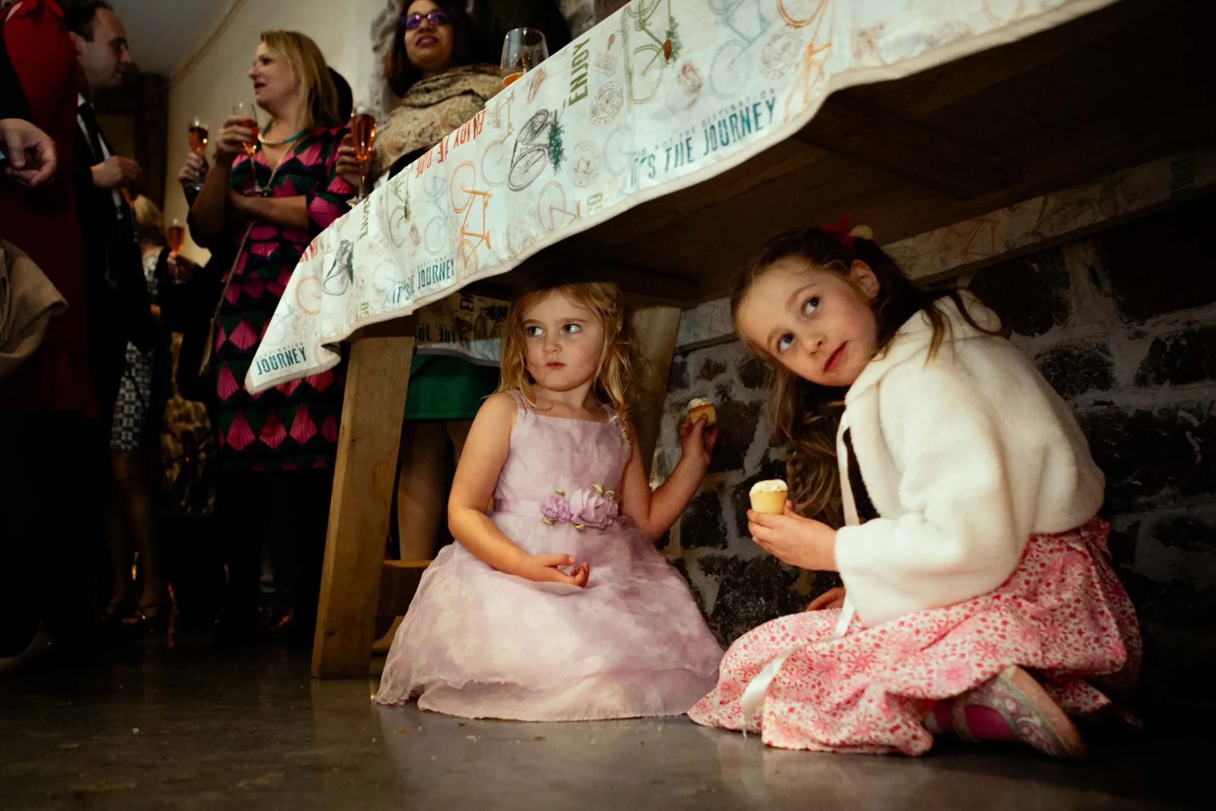 Two young girls are sitting under a table at a party, eating cupcakes, while adults are standing and chatting in the background.