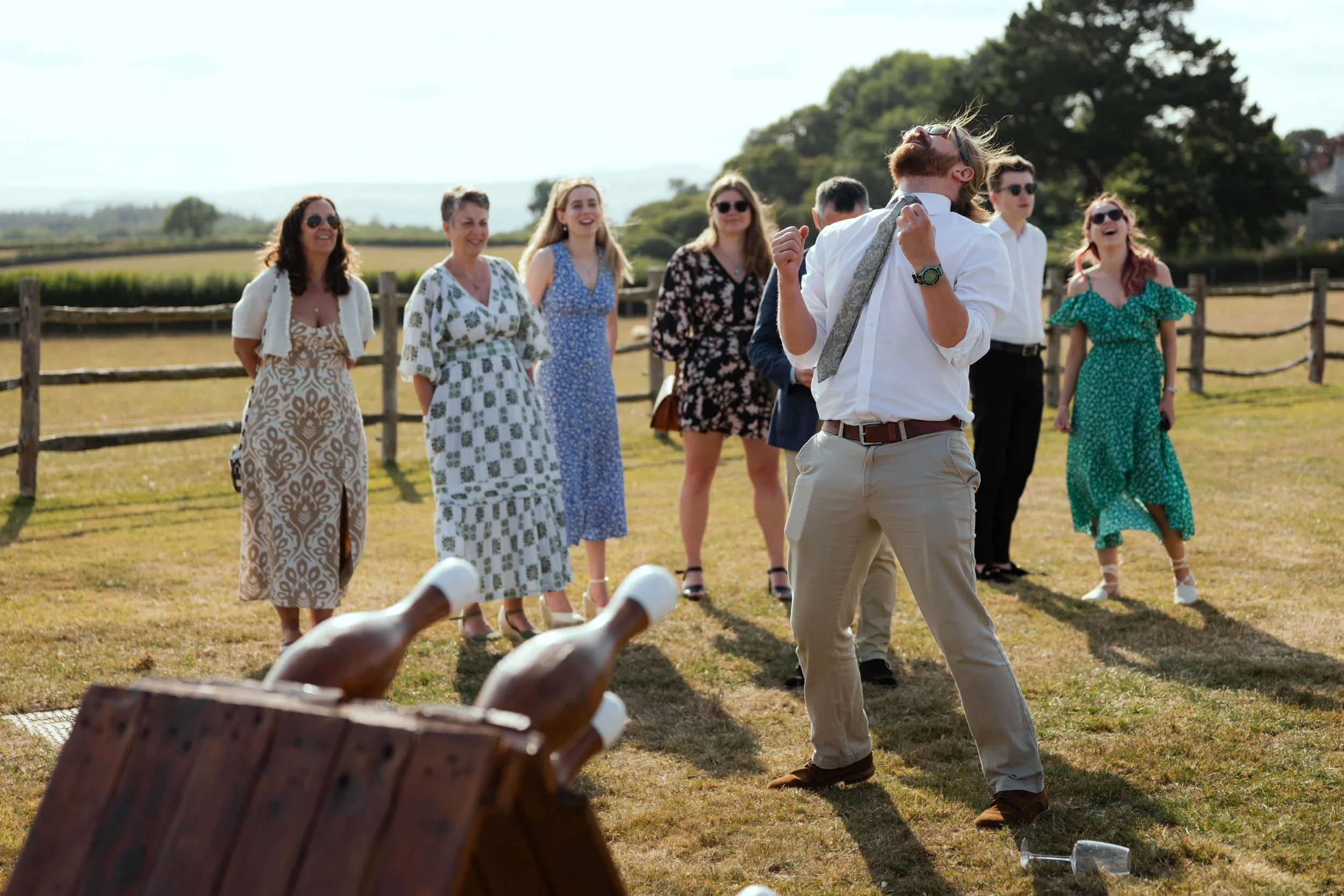 A man in a white shirt and beige pants dancing at an outdoor gathering with a group of women in summer dresses watching and smiling. There are bottles and a wine glass on the ground, and a wooden fence with trees in the background.