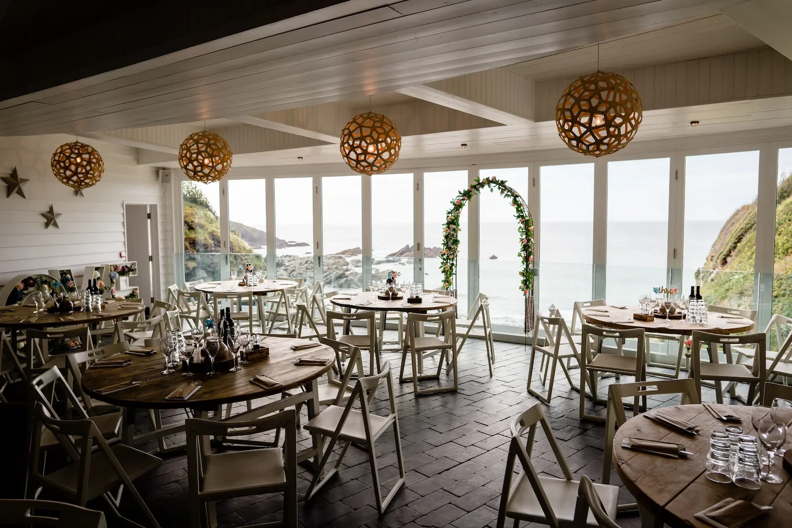 Beachside restaurant with round tables set with glassware and napkins, decorated for a wedding with a floral arch, overlooking the ocean through large windows, with hanging decorative lights.