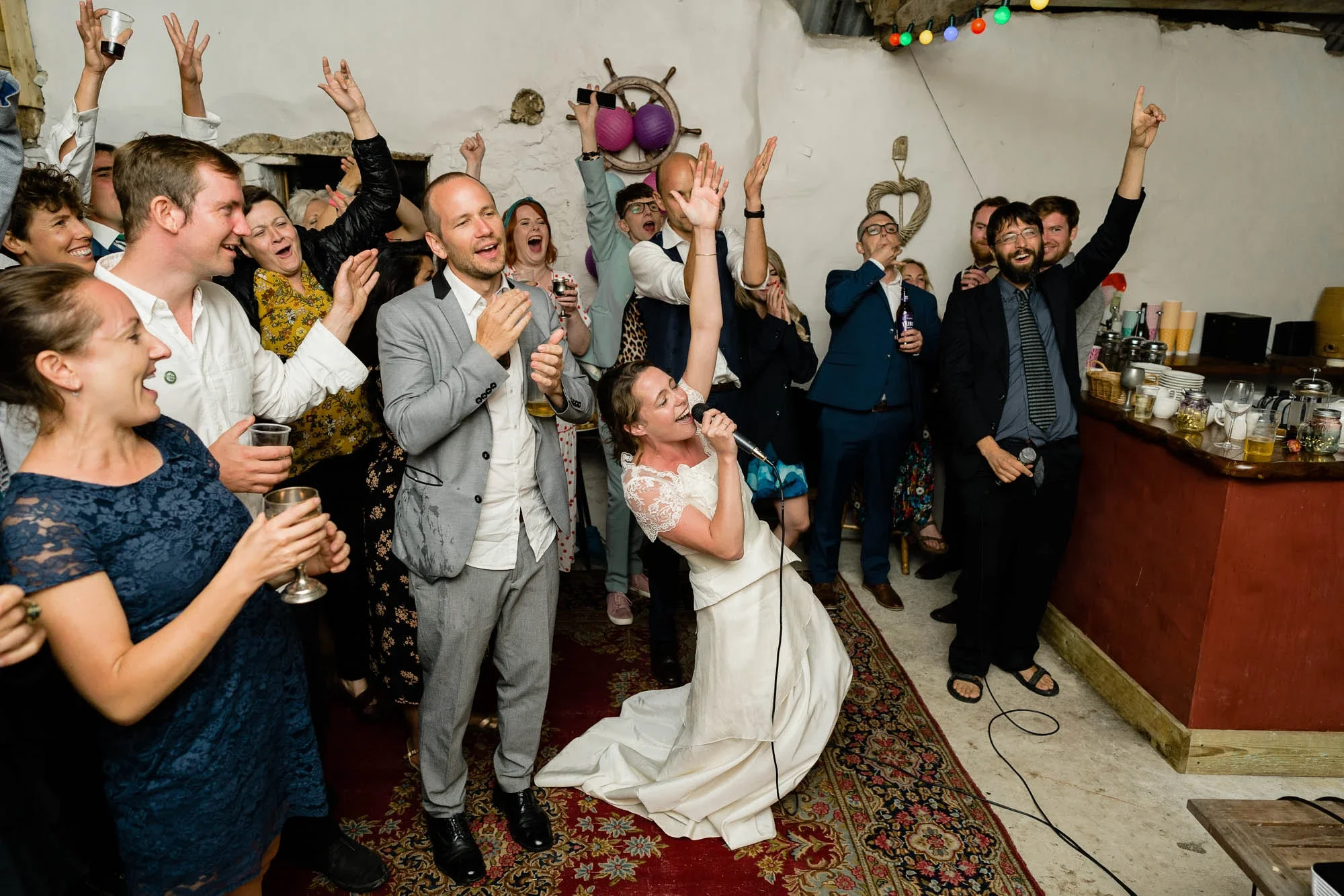 Group of people celebrating at a wedding reception, with the bride singing into a microphone, surrounded by guests dancing and holding drinks, in a decorated indoor setting.