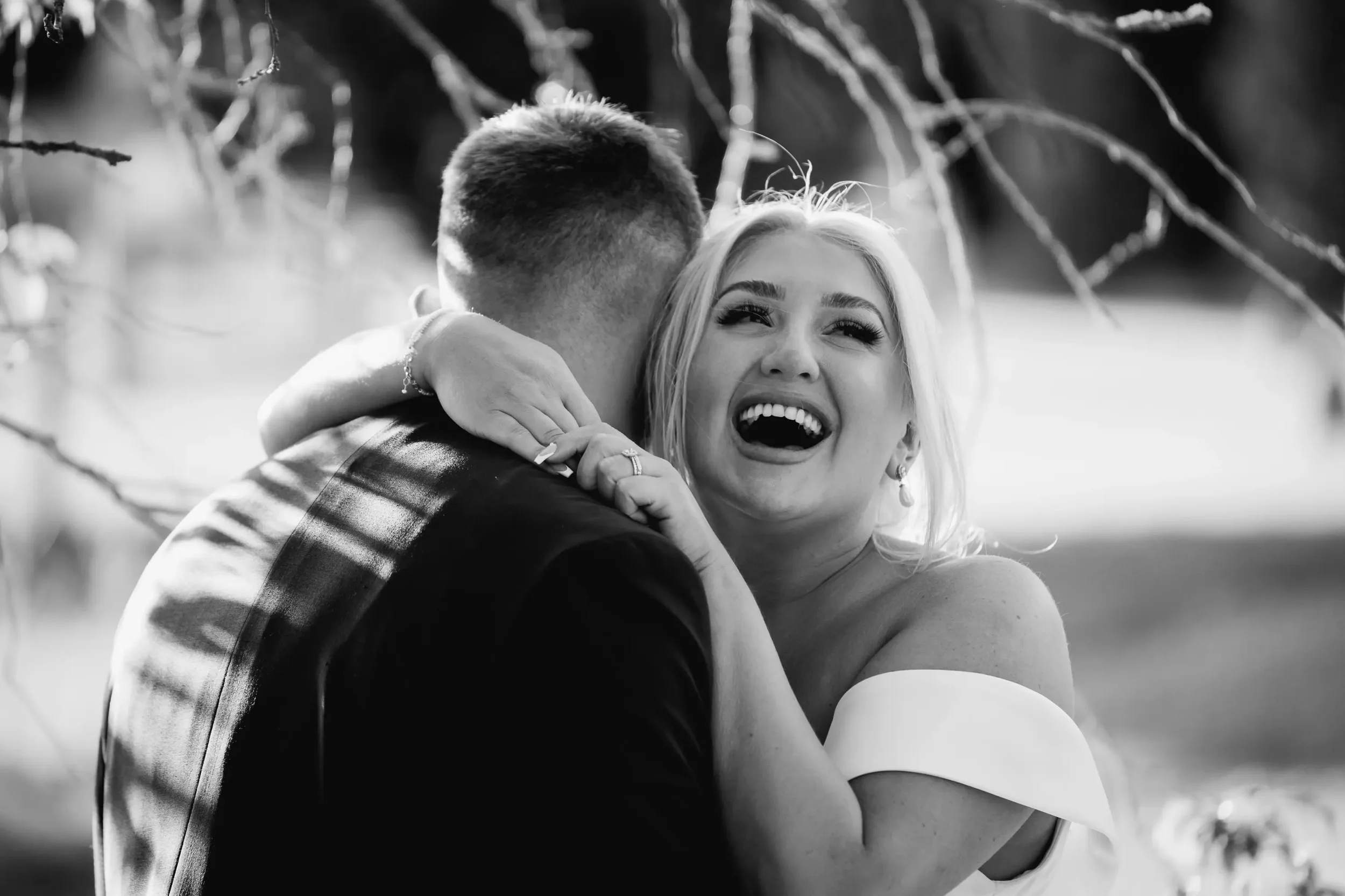 A joyful bride and groom embrace outdoors, with the bride smiling and laughing while hugging the groom.