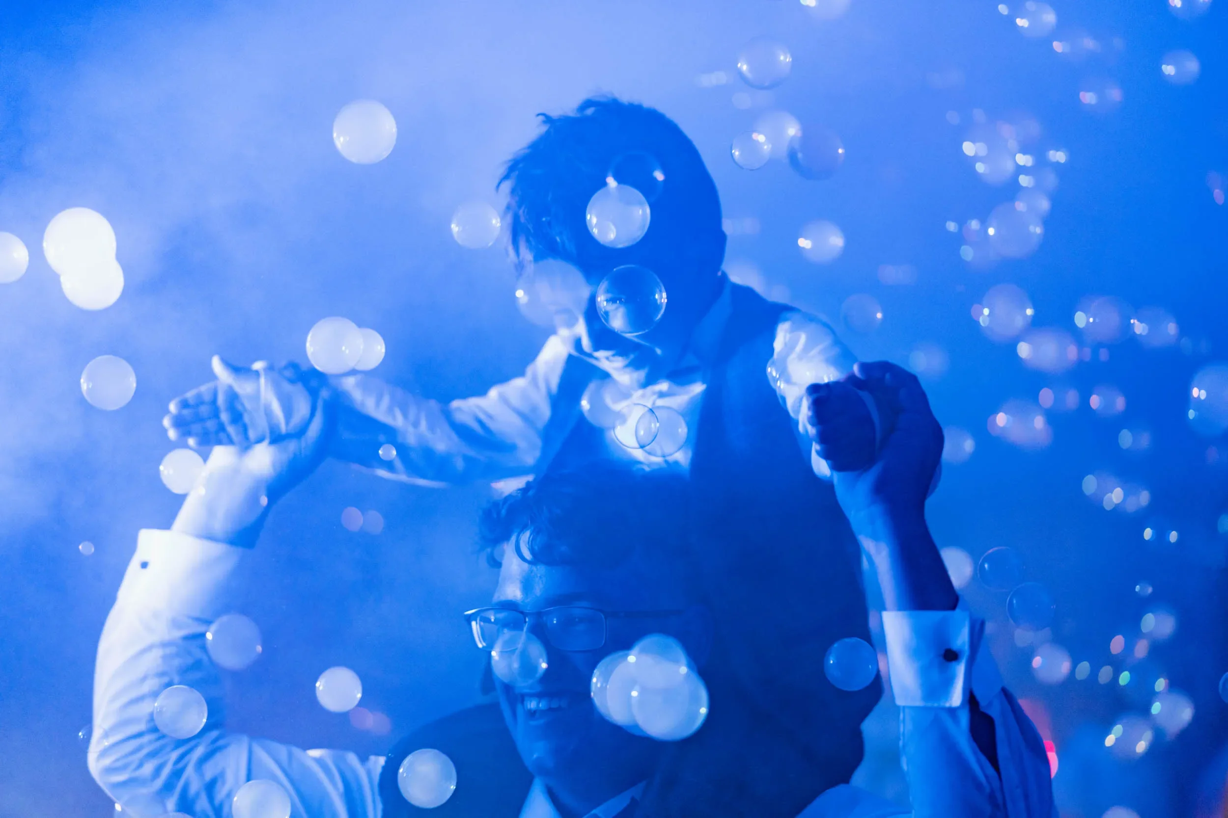 Two men in formal shirts, one wearing glasses, are playing in a blue-toned room filled with floating bubbles. One man is on the other's shoulders, with arms outstretched and holding hands, smiling and appearing joyful.